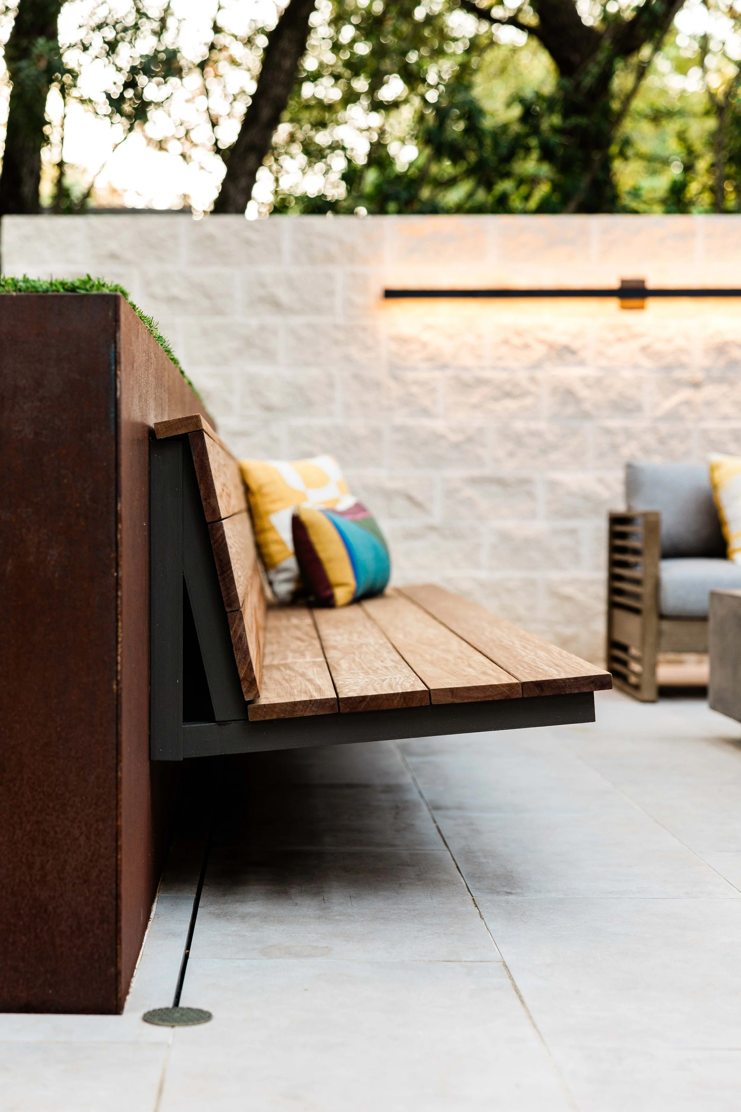 Outdoor seating area with a wooden bench with colorful pillows, a concrete floor, and a beige stone wall with a horizontal light fixture, surrounded by trees.
