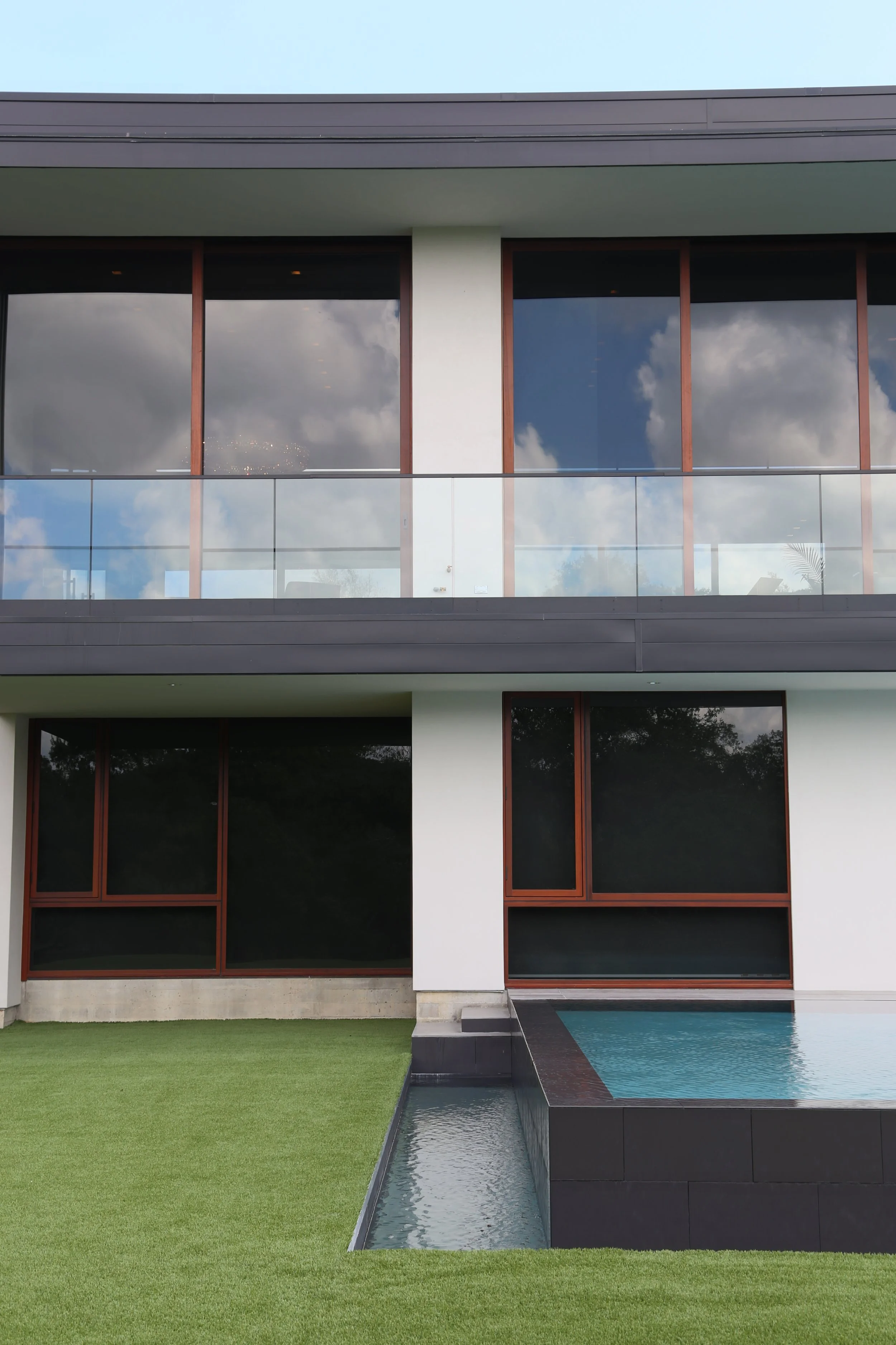 Modernist architecture meets serene luxury—this rear view captures a minimalist white and black facade reflected in a serene pool, framed by clean lines, dark-framed glass windows, and turf lawn
