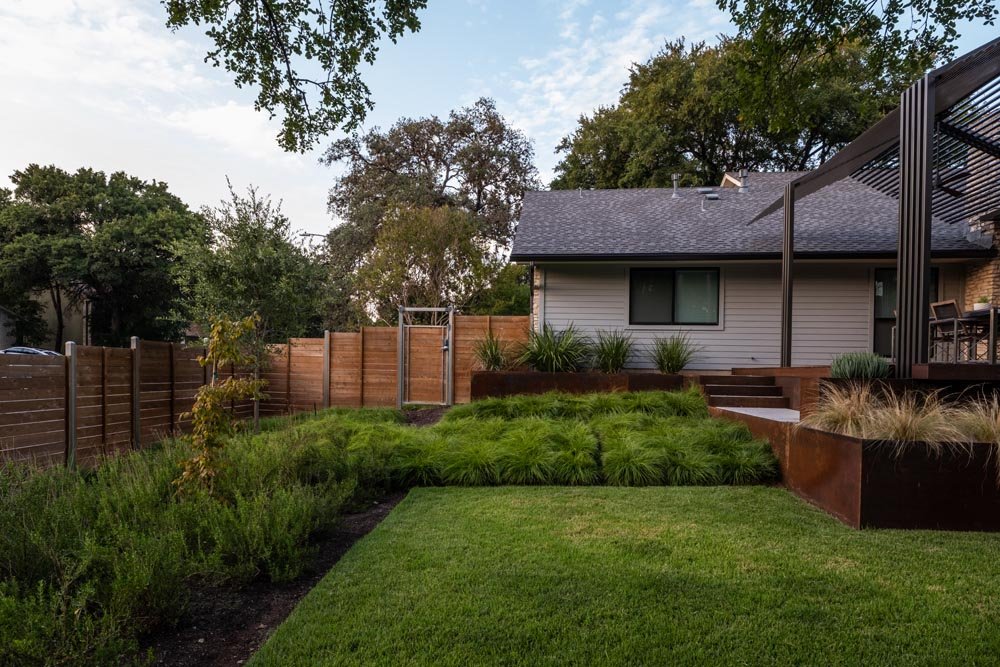 View of a modern backyard with native grass lawn and cedar privacy fencing, framed by layered retaining walls and lush plantings