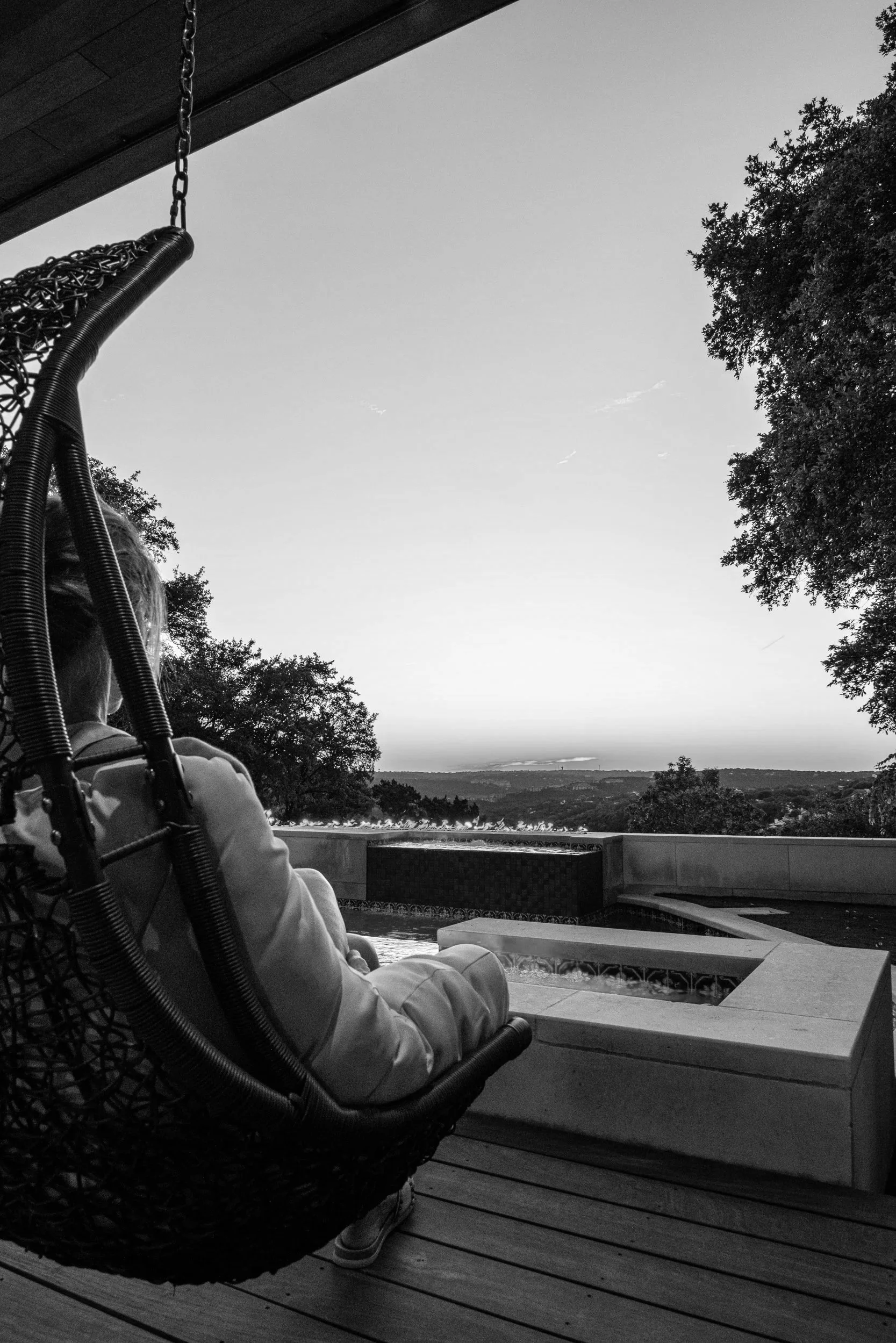 Person sitting in a woven chair on a wooden deck, overlooking a landscape with trees and a horizon, with a water feature in front.