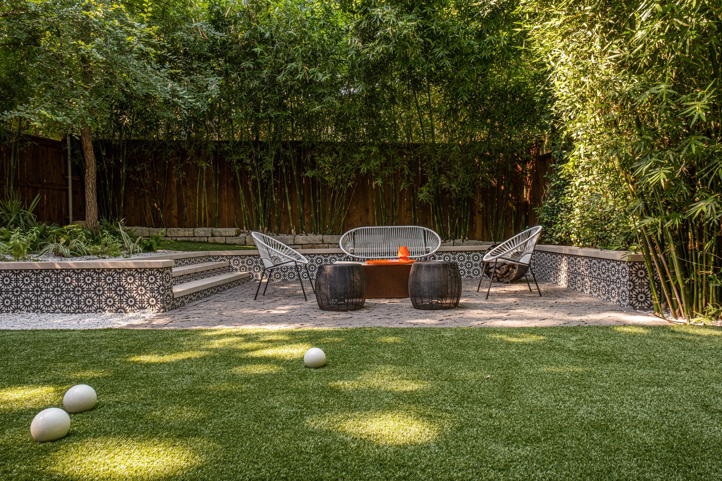 Low-angle view of a bocce ball court with a synthetic turf lawn surrounded by bamboo, patterned tile walls, and mid-century modern patio seating in the background. A cluster of white bocce balls is scattered in the foreground.