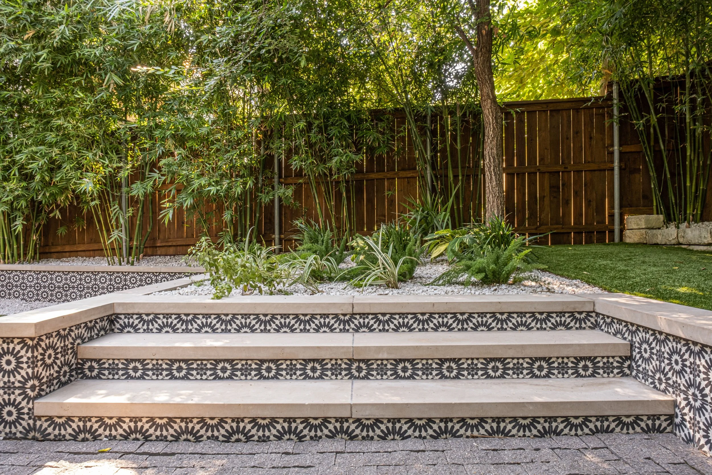 Decorative concrete tile stair risers with smooth limestone treads create a bold yet refined transition between gravel patio and elevated synthetic turf area. Bamboo and layered shade plants soften the hardscape and provide a private, leafy backdrop