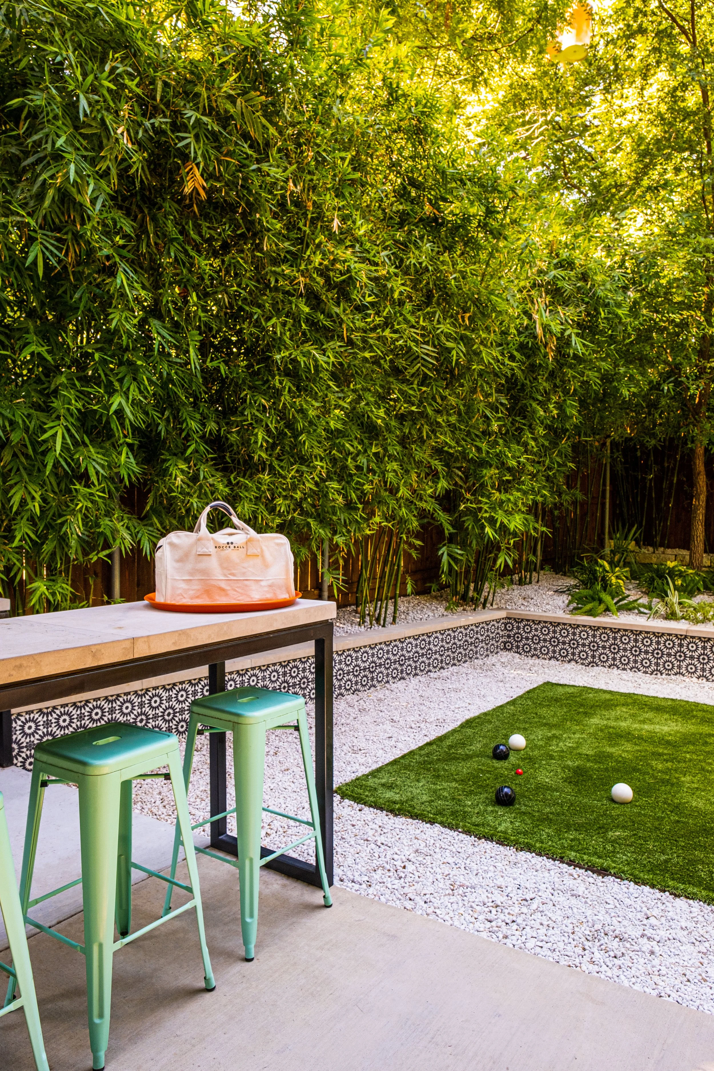 Outdoor bar overlooking a petite bocce court framed by encaustic tile planters, bamboo screening, and white gravel — a vibrant backyard retreat for playful gatherings and slow afternoons