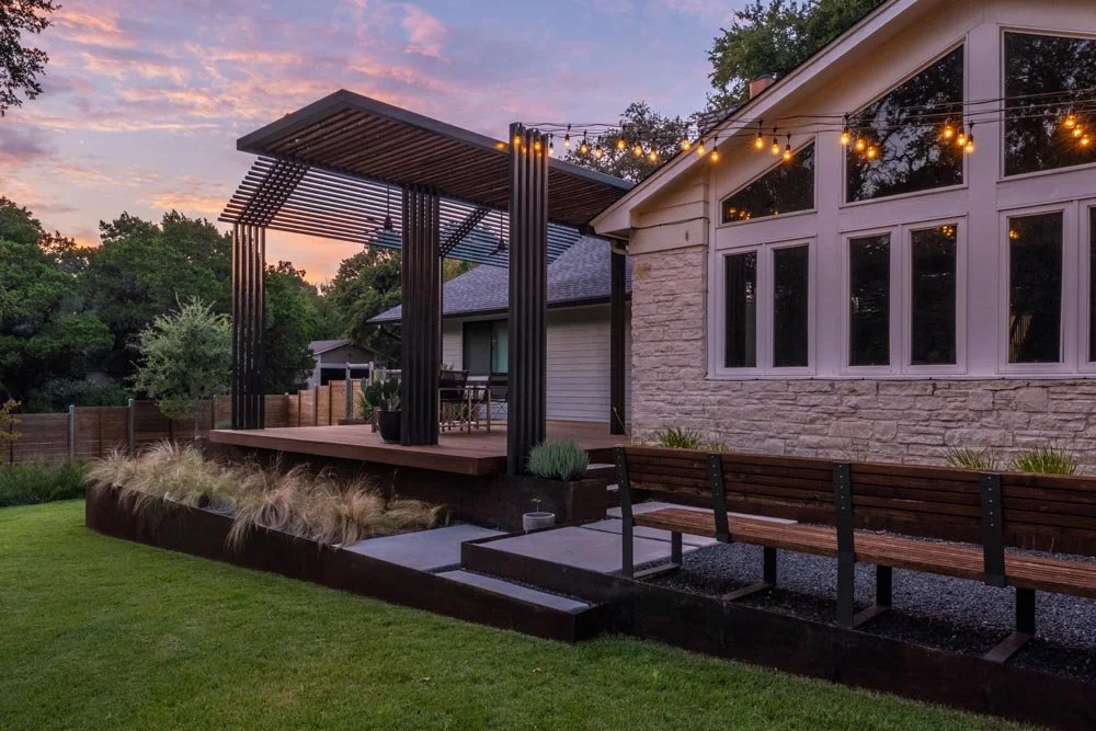 Evening view of the custom louvered steel pergola casting rhythmic shadows over a dining patio adjacent to the home’s stone exterior