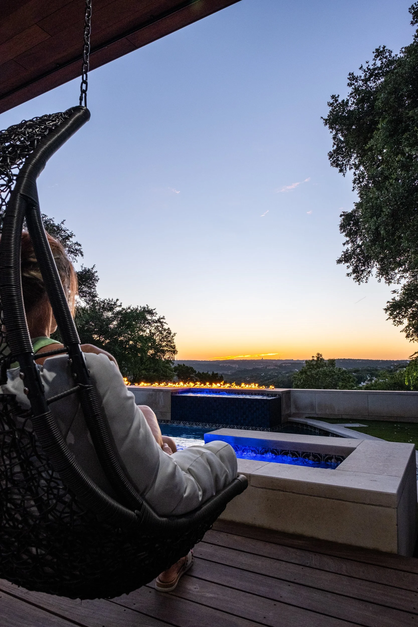 Person sitting in a hanging chair on a patio, looking at a sunset over a landscape with trees and distant city lights, near a pool with blue lighting.