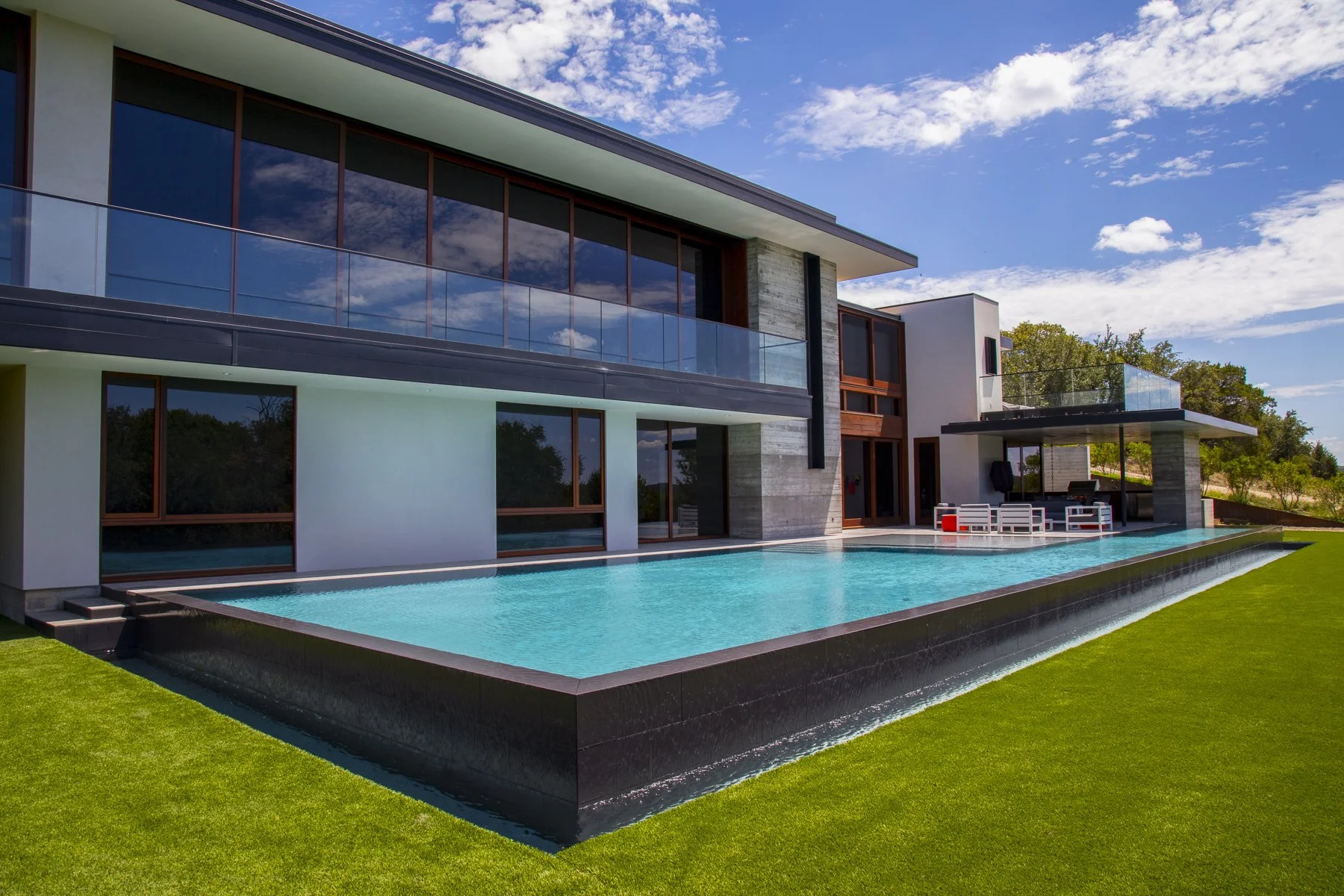 Modern hilltop home with a sleek glass façade and negative-edge pool that mirrors the sky, anchored by a vibrant turf lawn and bold architectural lines