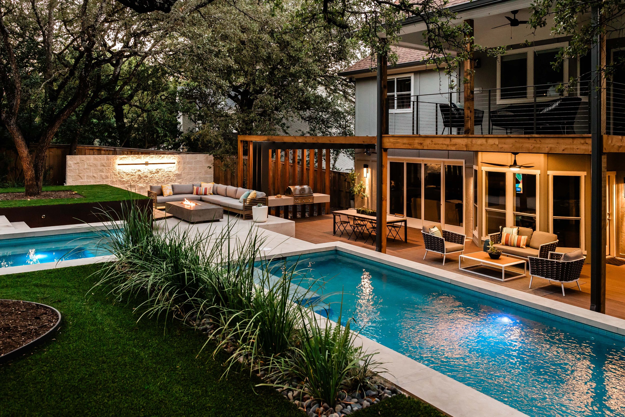 Evening view of a contemporary backyard with glowing pool lights, expansive glass doors, and a warm colored composite deck outdoor living space