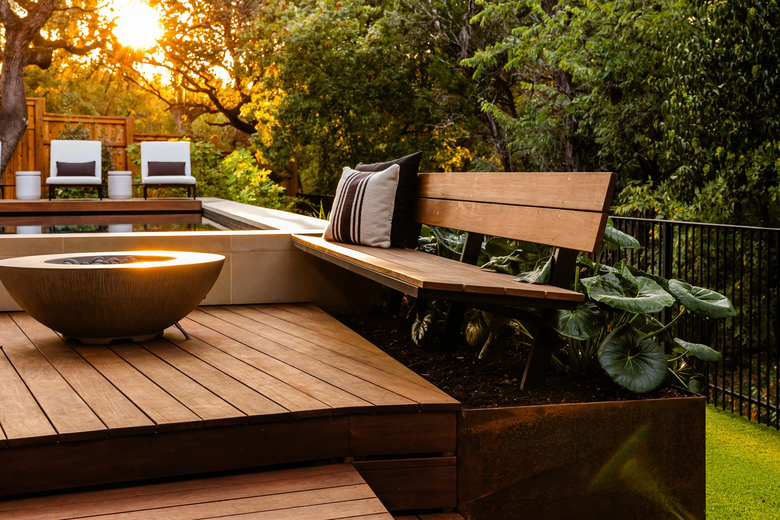 Wooden bench and fire bowl on a warm-toned deck by poo surrounded by lush foliage at sunset