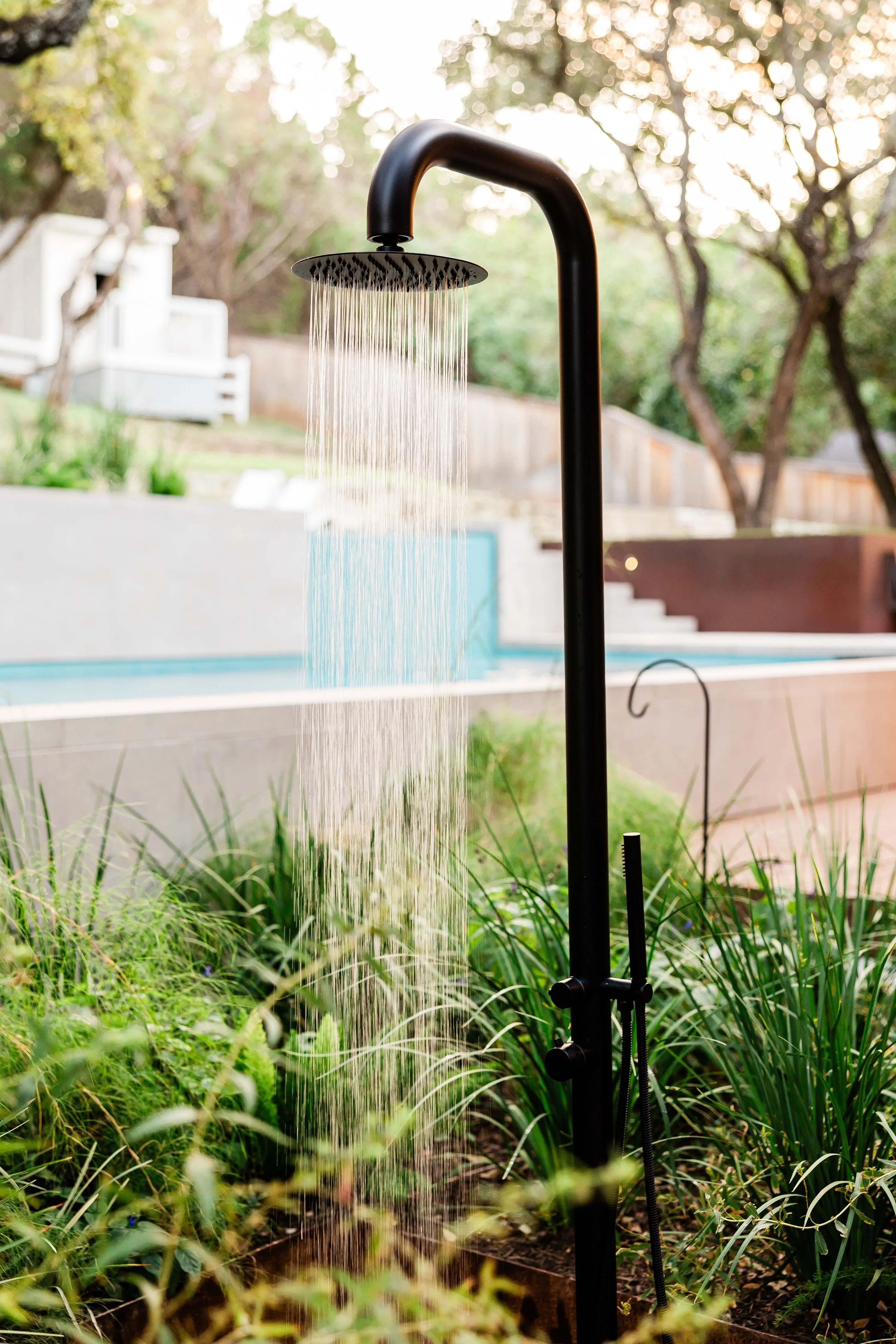 A sleek, black outdoor shower stands amid lush ornamental grasses, with water streaming down in front of a clean-lined modern pool and layered retaining walls