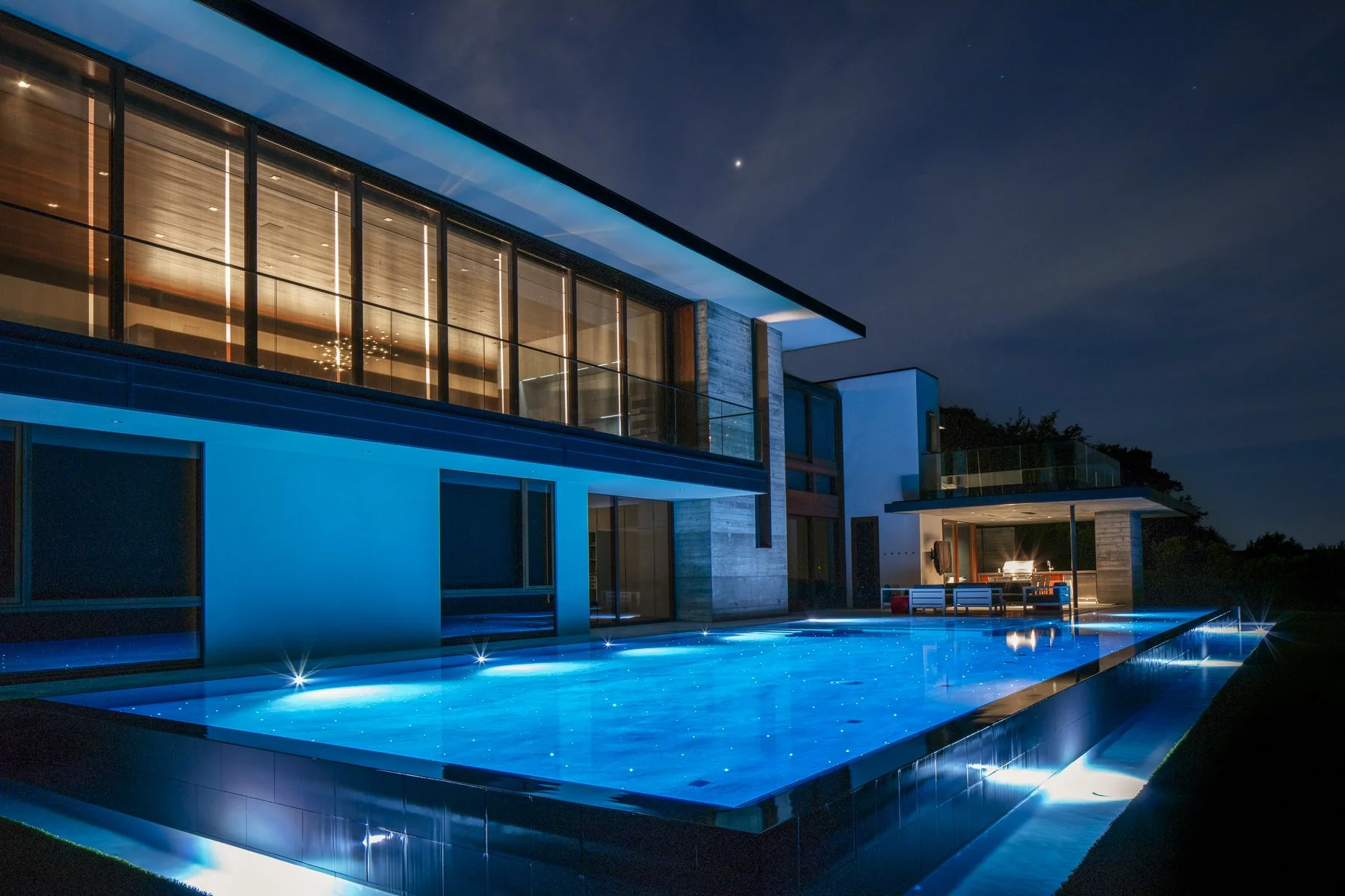 Evening view of a luxury modern home with floor-to-ceiling windows reflecting warm interior light, beside a glowing blue negative-edge pool that mirrors the night sky