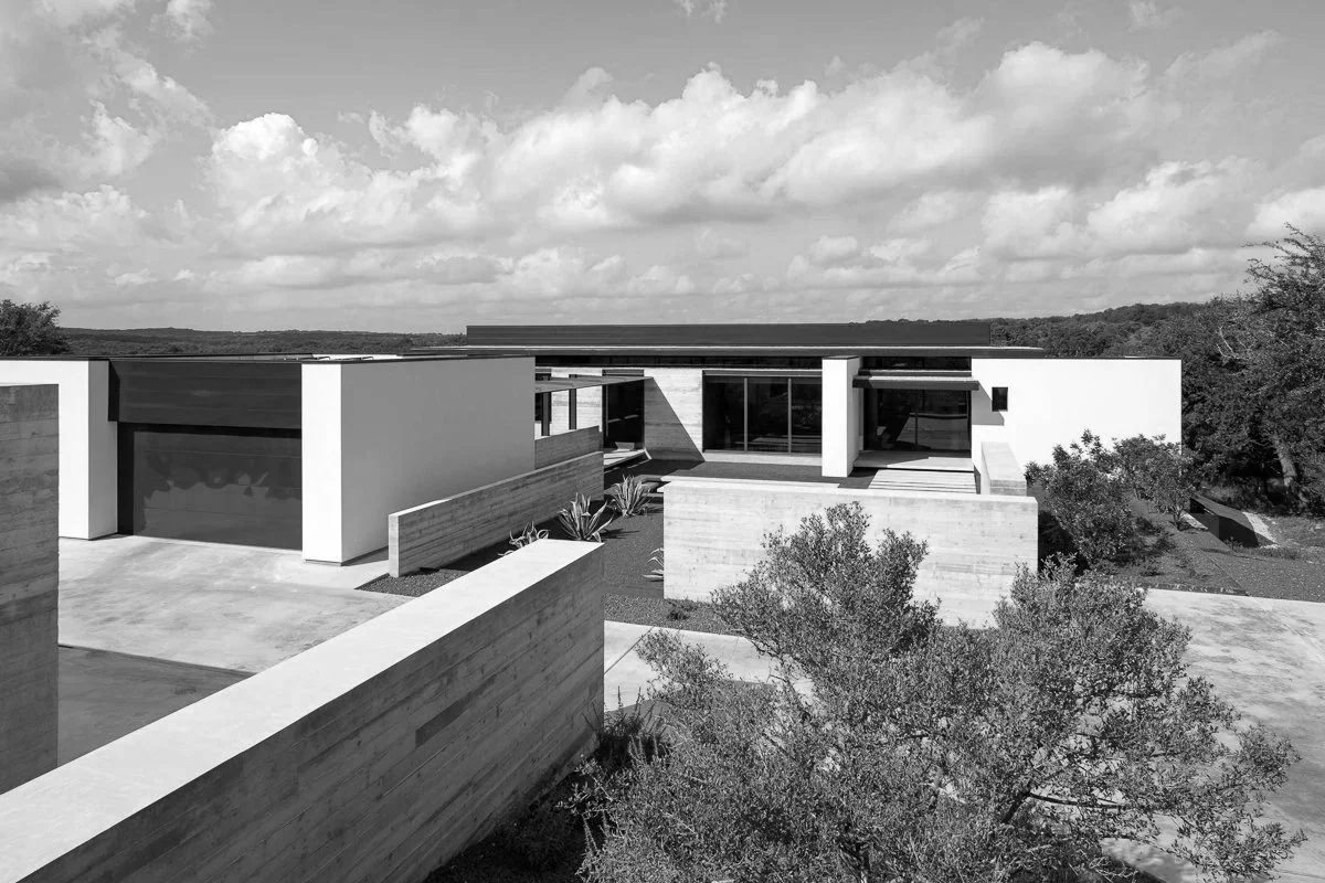 A modern minimalist house with flat roofs, large glass windows, and clean lines, surrounded by trees and a concrete driveway, under a cloudy sky.