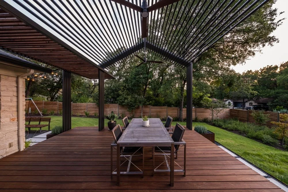 A modern outdoor dining area with a slatted steel pergola overhead, casting dramatic shadows on a wooden deck. The setting features a sleek rectangular dining table with black chairs and a view of a lush backyard enclosed by a horizontal wood fence. 
