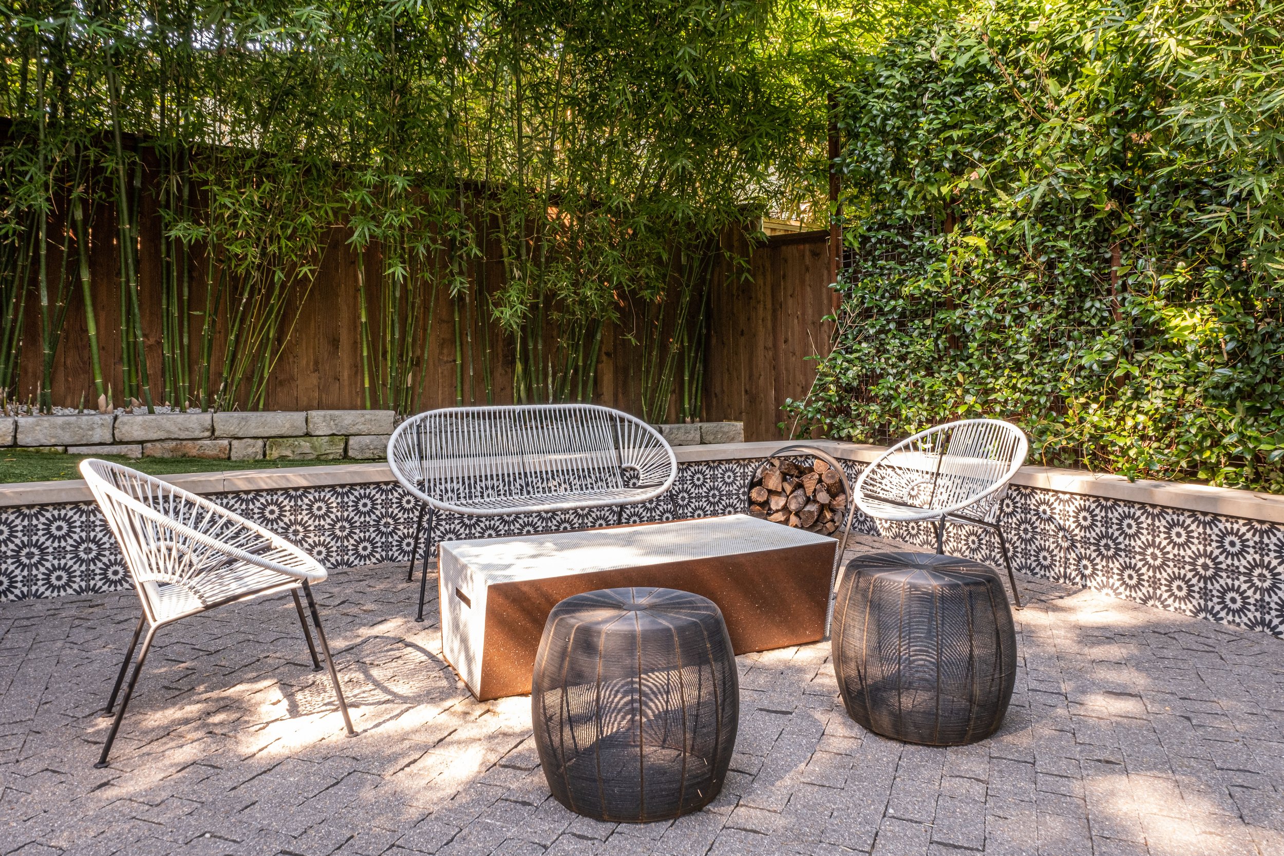 Cozy outdoor fire pit lounge set within a bamboo-lined backyard, featuring a modern woven loveseat, two minimalist wire chairs, and two sculptural black metal stools around a custom geometric fire pit table, with decorative tile-faced seating walls c