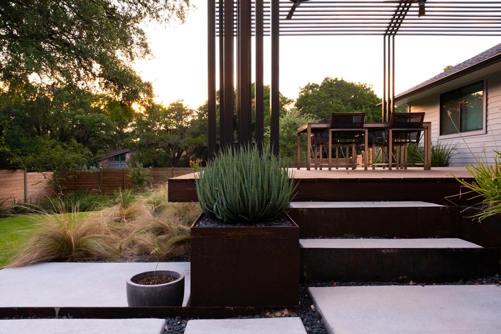 Close-up of minimalist concrete stairs framed by steel planters and grasses, leading up to a dining deck shaded by a linear steel pergola. The warm sunset light highlights the crisp material contrasts