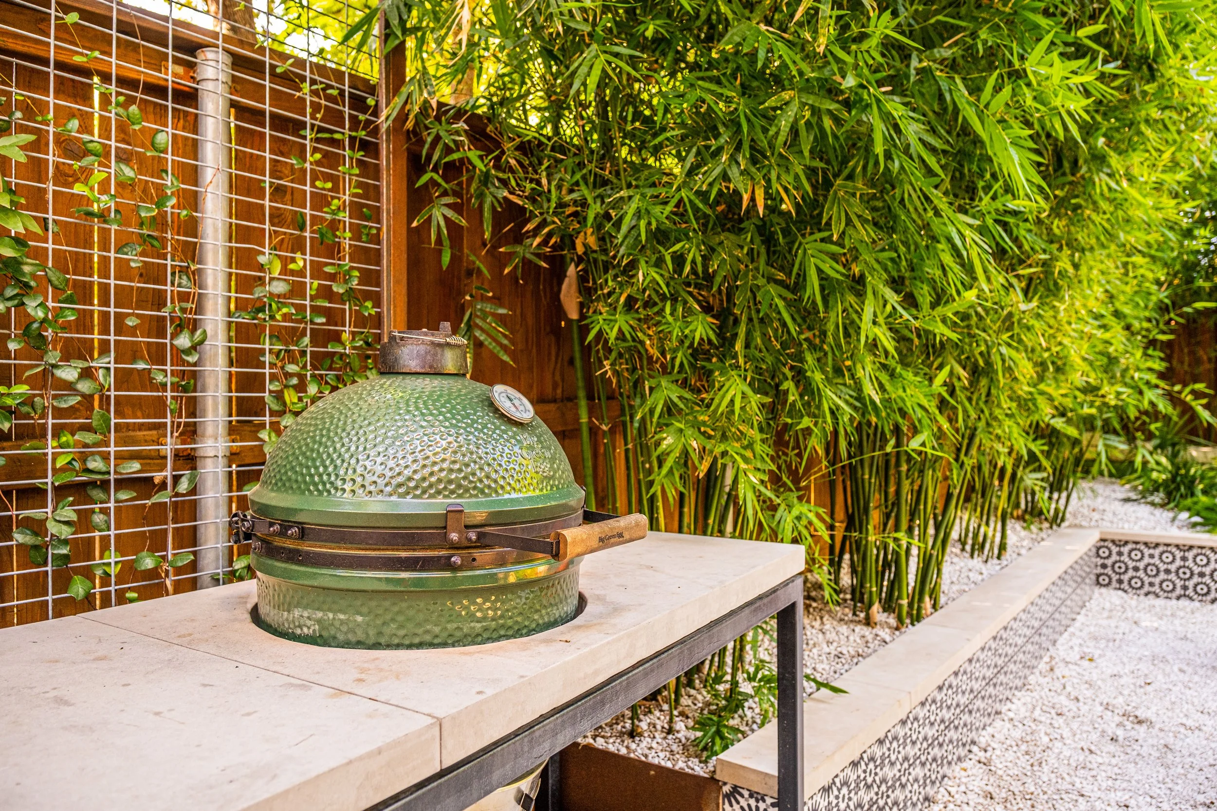 Green ceramic Big Green Egg on a stone countertop framed by lush bamboo and white gravel, nestled in a modern urban backyard oasis