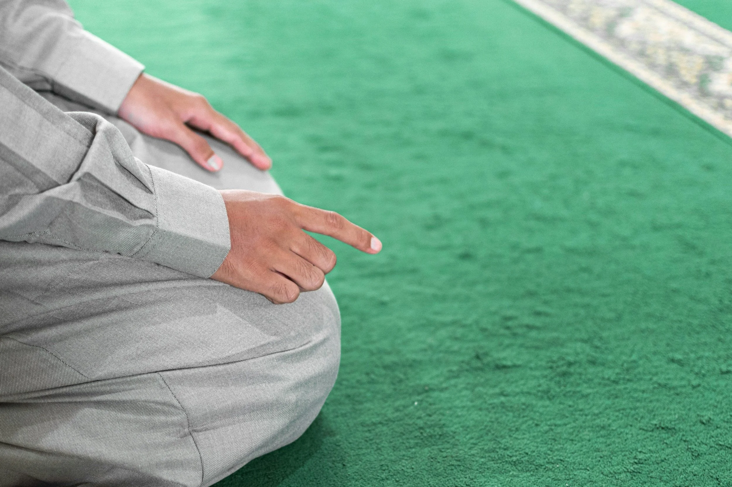 Person dressed in light gray clothing kneeling on a green prayer rug, with hands resting on the knees, pointing downward with index fingers.