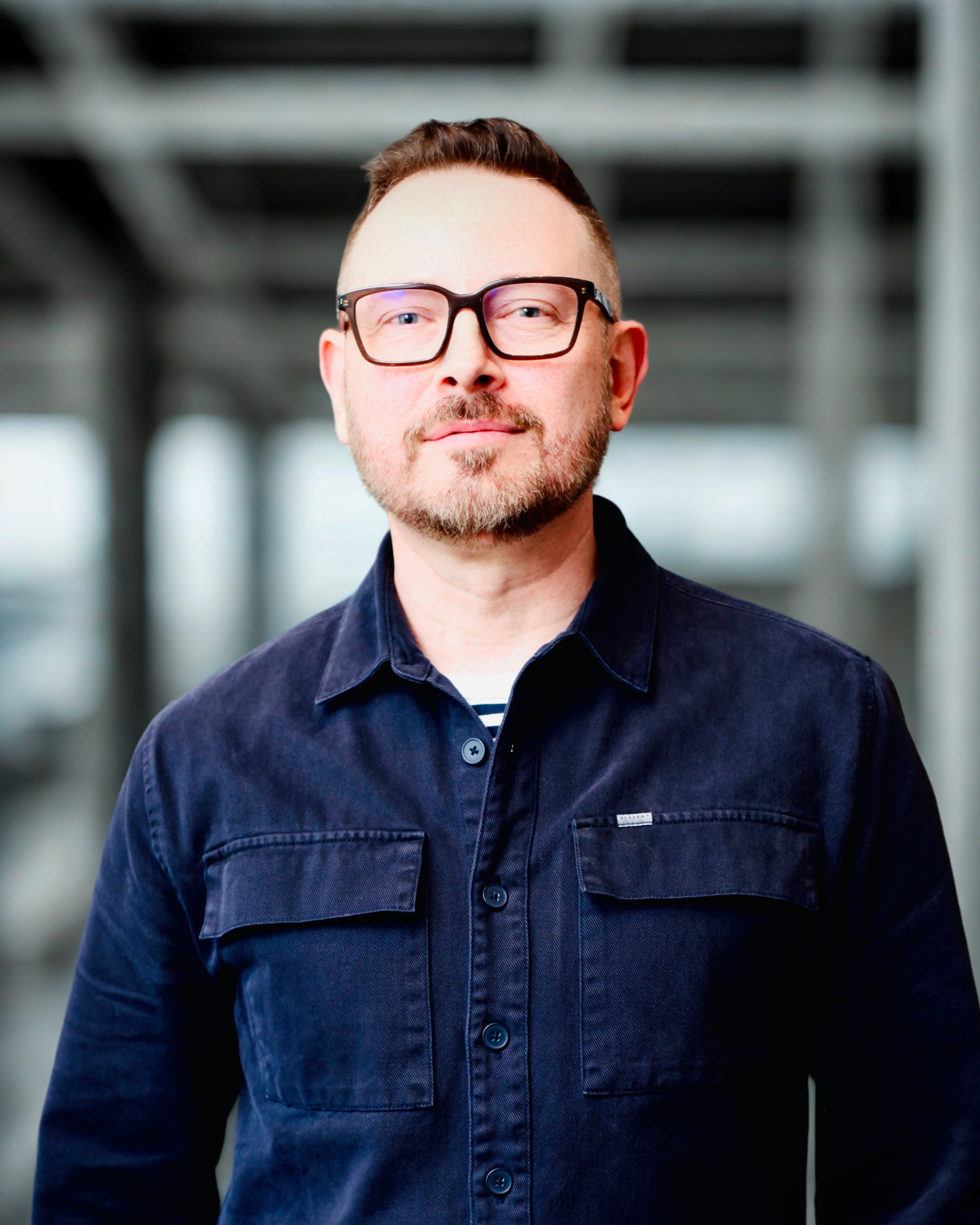 A man with short hair, glasses, and a beard wearing a dark denim shirt standing outdoors with a blurred industrial background.