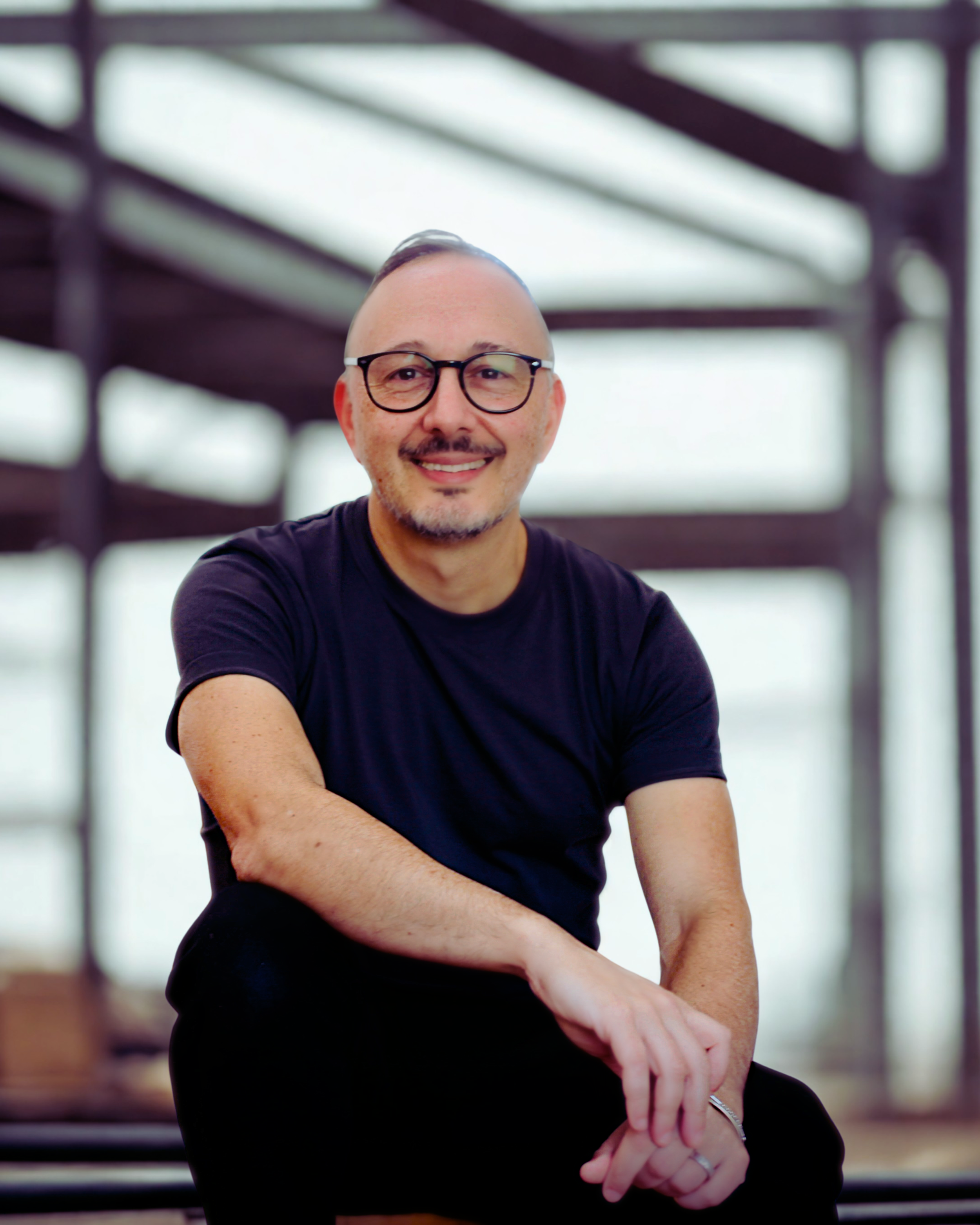 A man with glasses and a black T-shirt, smiling and sitting with crossed arms in a modern building with large windows.