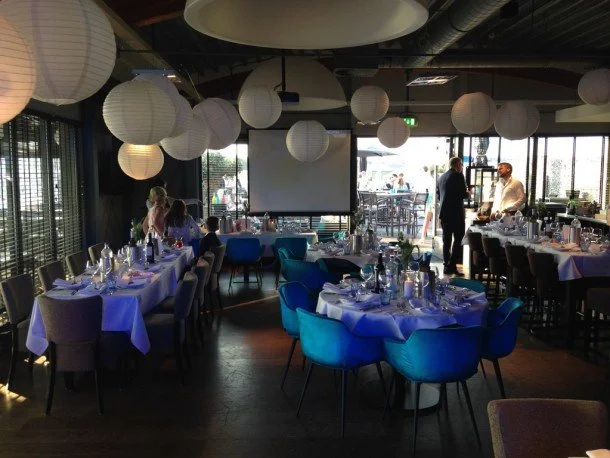 restaurant De Dining met ronde papieren lampenkappen aan het plafond, tafels met wit tafelkleed en glazen, en mensen die praten en wachten.
