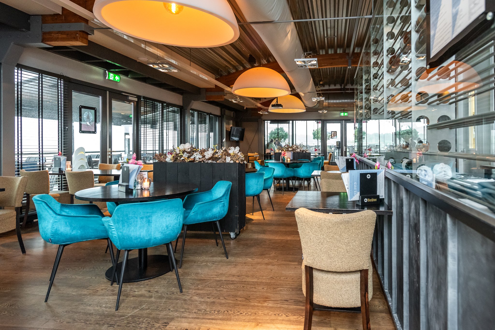 Interieur van een modern restaurant De Dining met houten vloeren, kleurrijke stoelen, grote hanglampen en grote ramen met luiken.