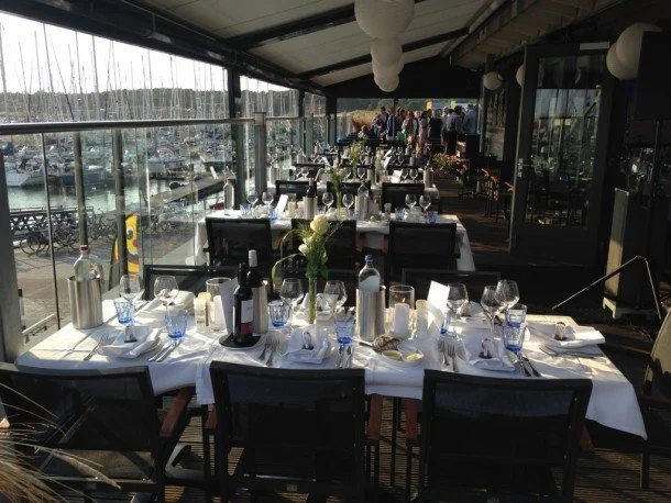 restaurant De Dining met gedekte tafels, glazen, borden en bloemen, op een terras met uitzicht op de haven van Vlieland vol boten.