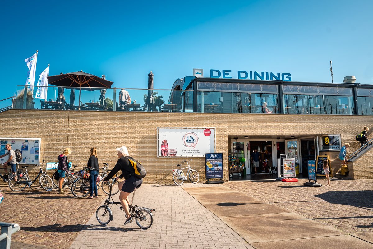 Buitenruimte met mensen, fietsen en restaurant De Dining met terras, onder een blauwe hemel.