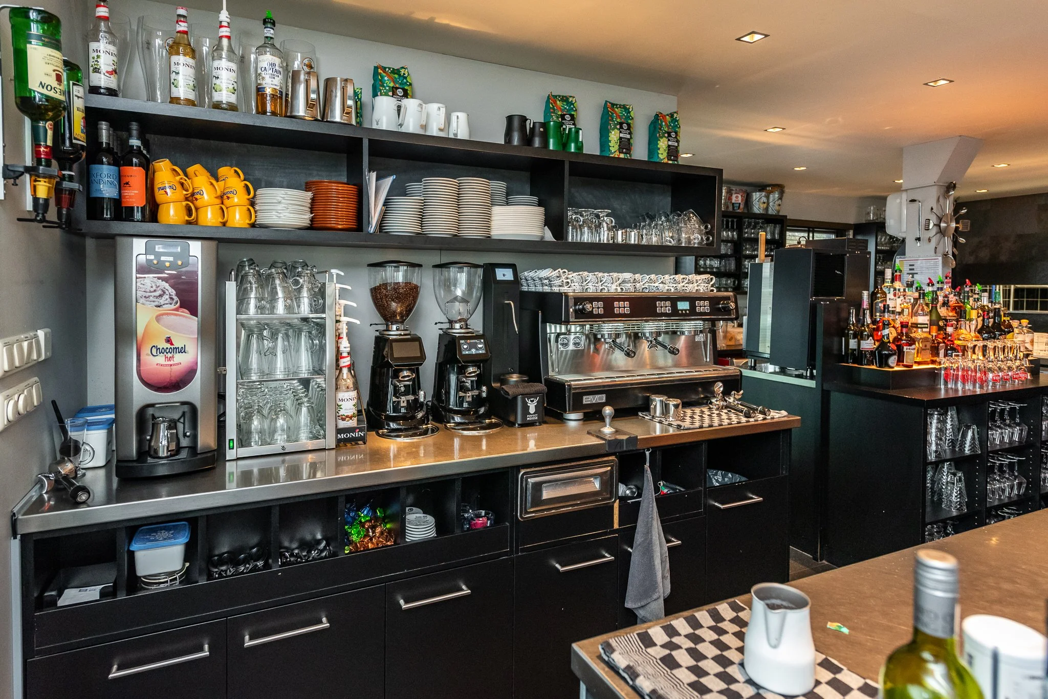 Een horecabar met koffiezetapparatuur, glazen, drankflessen en koffiemokken op de toonbank en schappen.