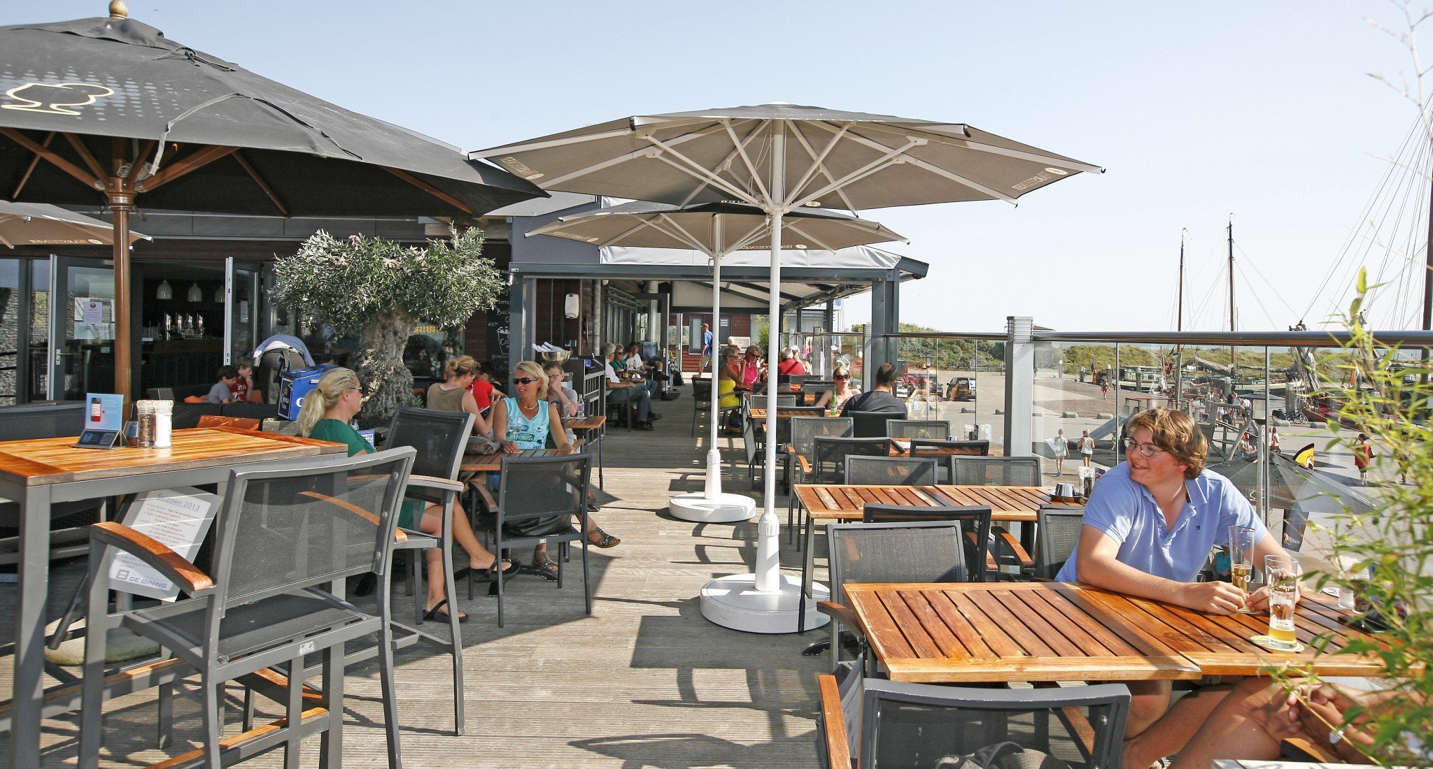 Buitenrestaurant De Dining aan het water met tafels, stoelen en parasols, mensen zitten te genieten van de zon en drinken.