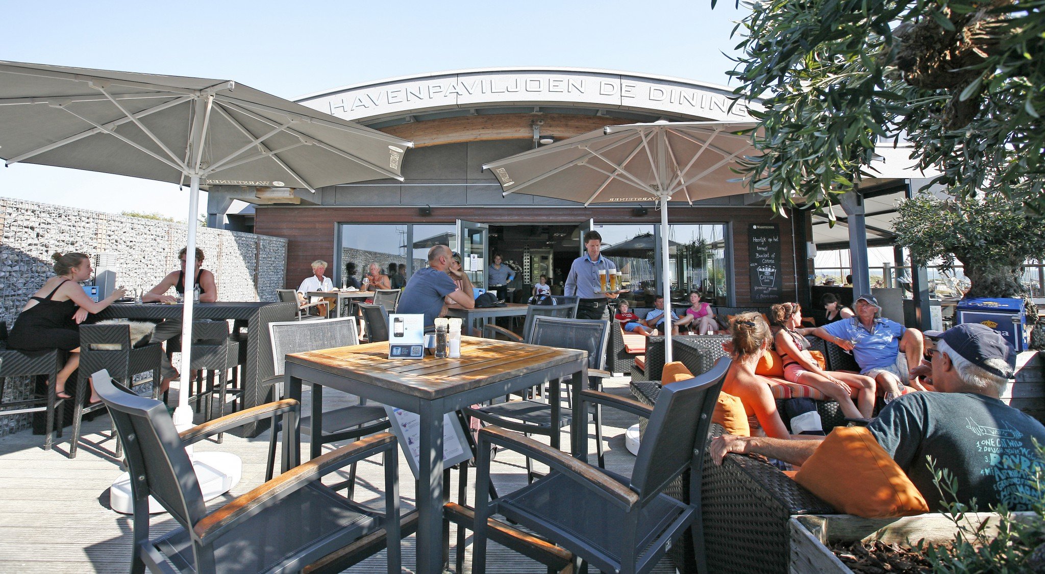 Buitenrestaurant De Dining met mensen die genieten van hun tijd, onder grote parasols, op terras met tafels en stoelen, en een gebouw met de tekst 'HAVENPAVILJOEN DE DINING' op de gevel.