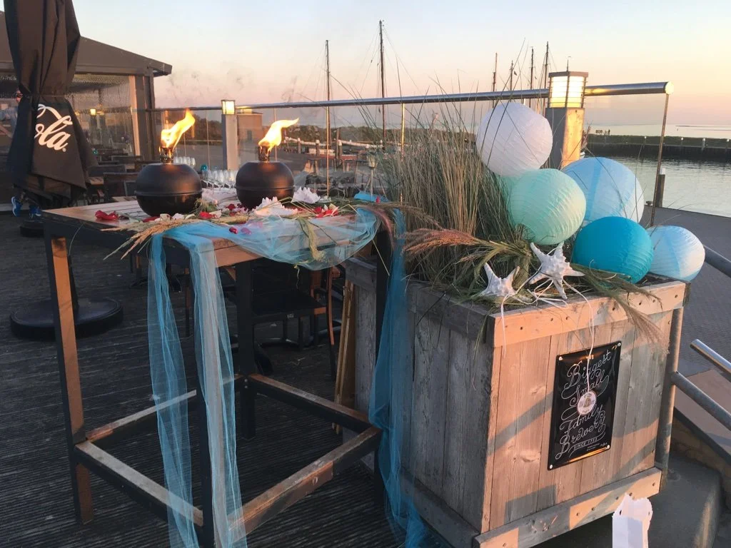 Decoratie op een terras met blauwe en witte papieren lantaarns, houten container met grassprietjes en sterrenvormige decoraties, twee bakken met vuurpotten, uitzicht op het water en schepen in de achtergrond.