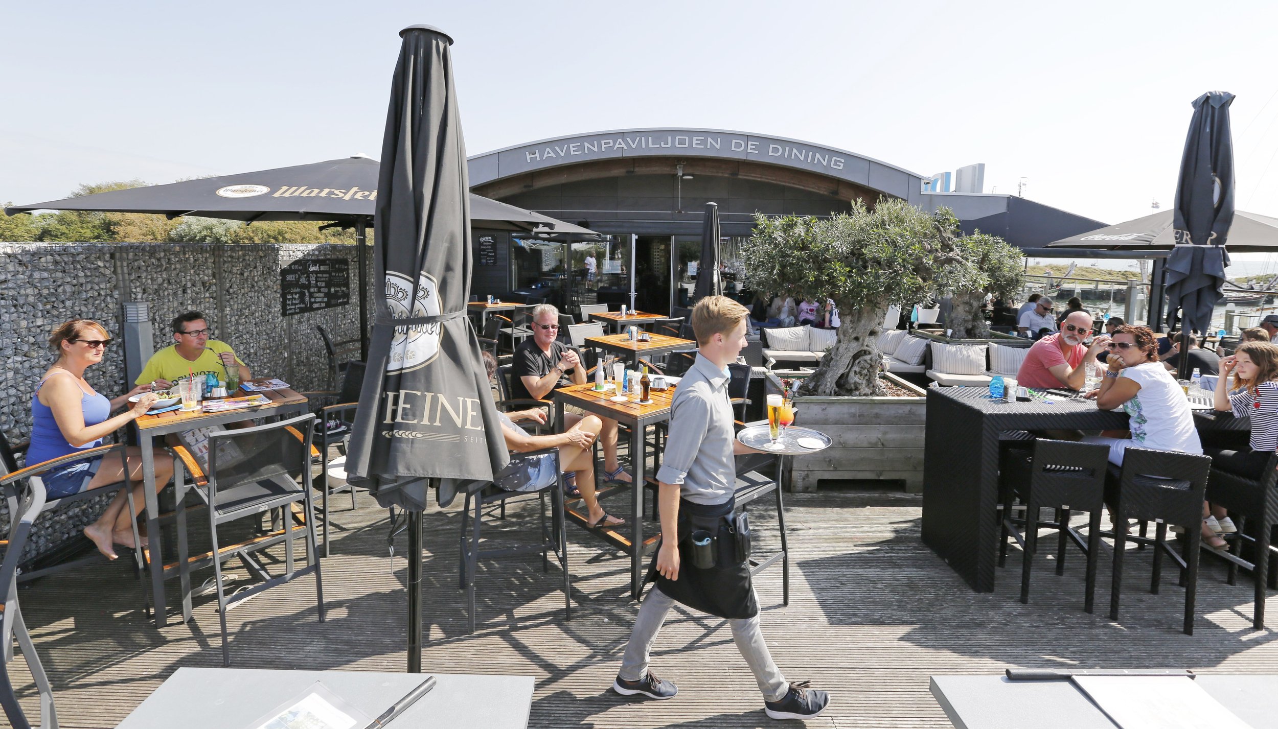Terras van De Dining met mensen die genieten van eten en drinken, sierboom in het midden, parasols met Heineken logo, gebouw met naam 'Havenpaviljoen de Dining' op de achtergrond, tijdens zonnige dag.