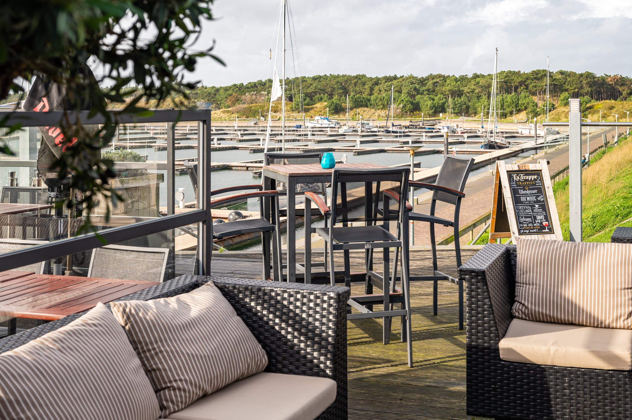 Uitzicht op een terras aan het water met buitenmeubilair en een haven van Vlieland met boten op de achtergrond.