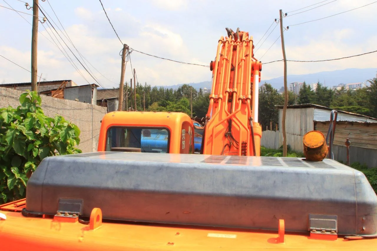 Farm construction vehicle with orange arm and metallic black cover on top, background shows wooden and metal sheds, power lines, trees, and distant hills.