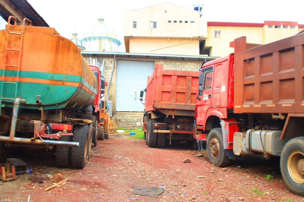Three large trucks parked on a dirt lot in front of a building with a blue-domed structure in the background.
