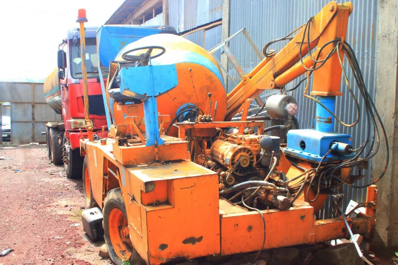 A mechanically operated concrete pump with a yellow arm and orange body, parked outdoors next to a corrugated metal wall.