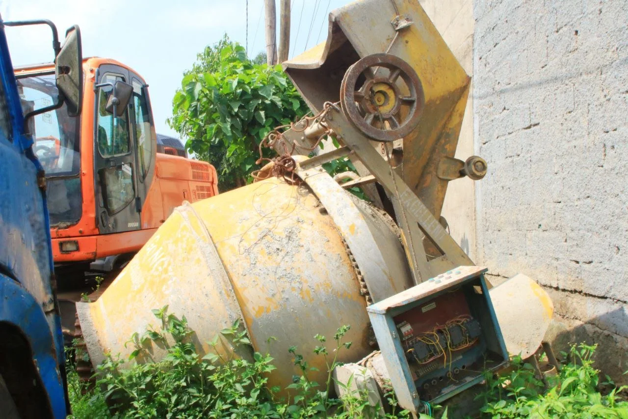 Old, rusted construction equipment lying on the ground near a concrete wall, with overgrown weeds and plants surrounding it, and an orange excavator in the background.