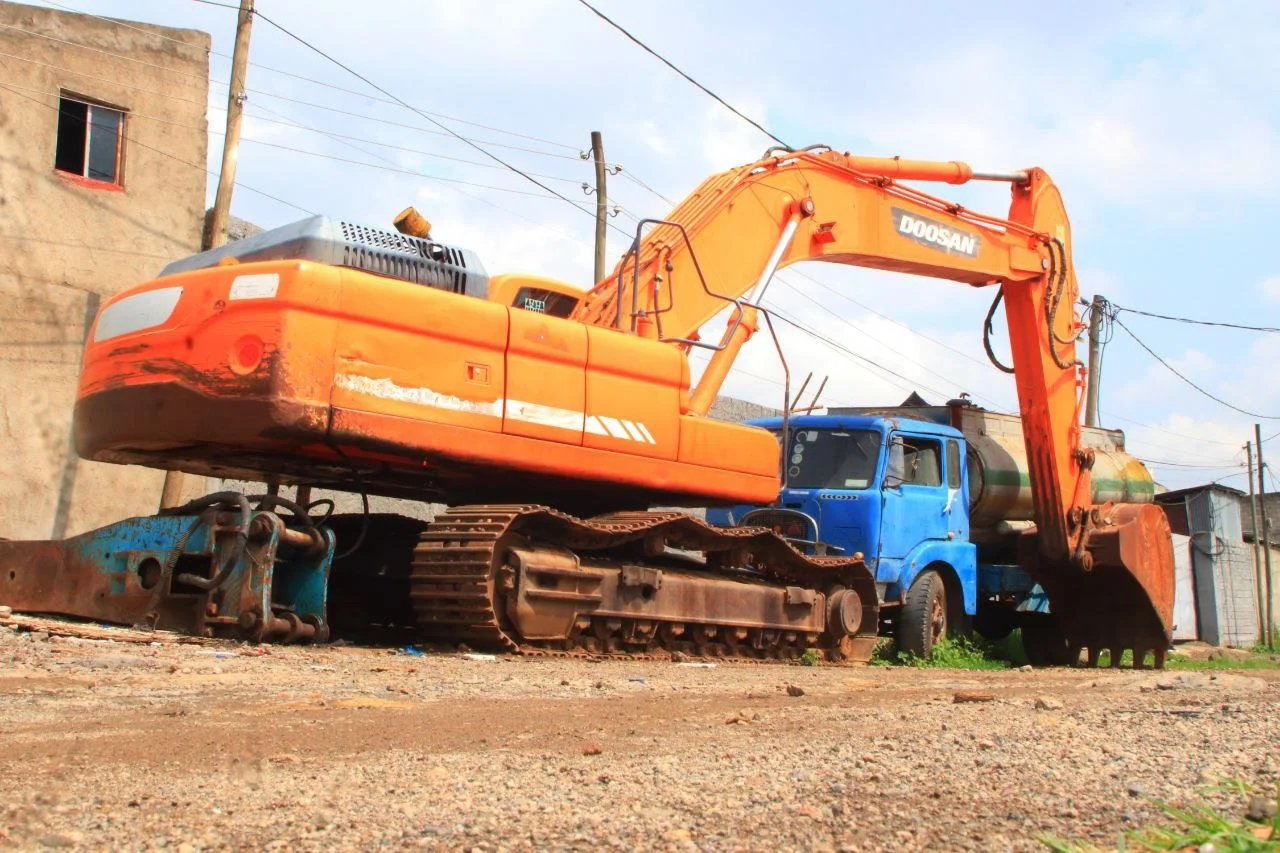 Orange Doosan excavator resting on the ground behind a blue truck in an outdoor setting with wires and buildings in the background.