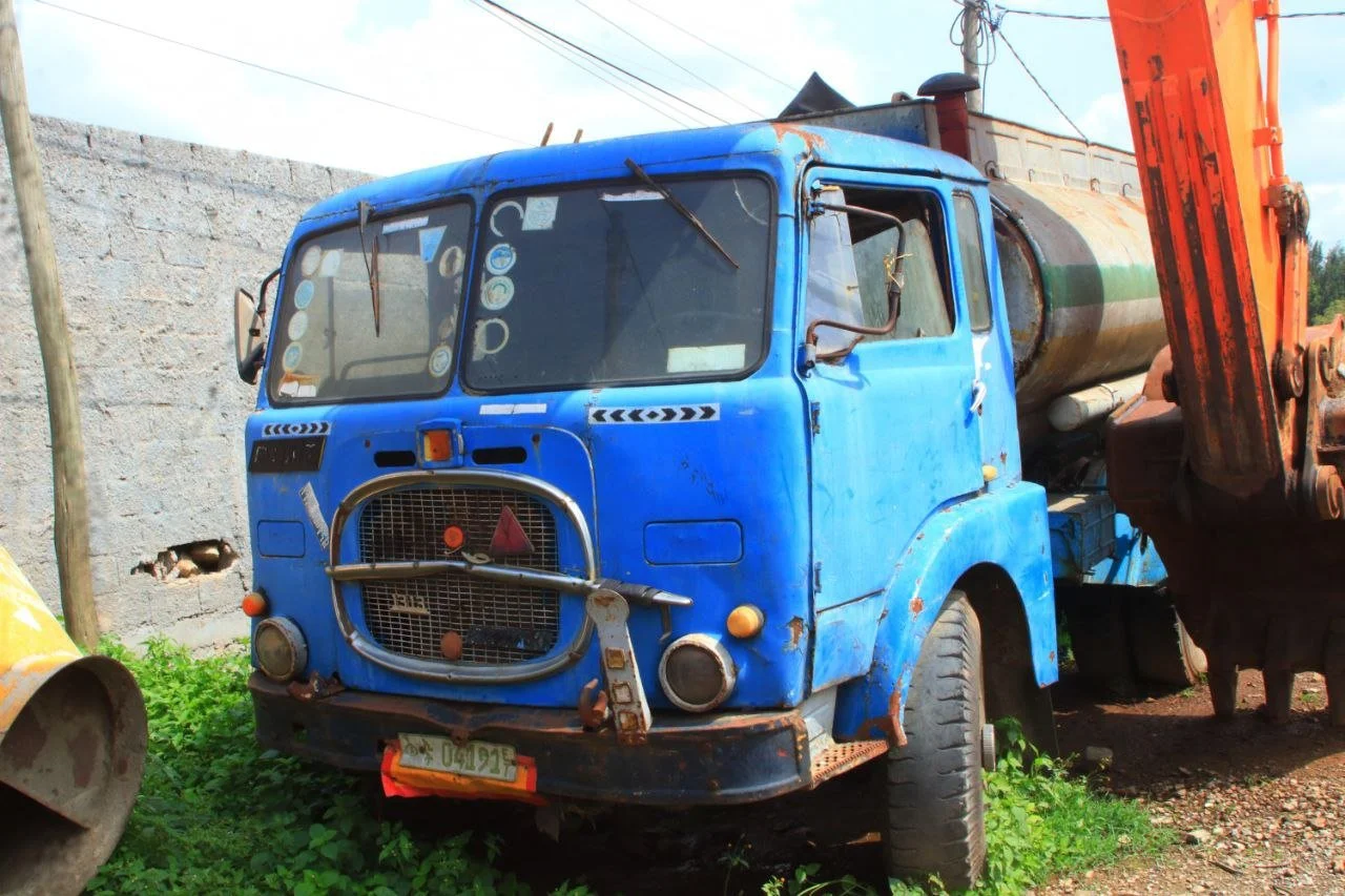 An old blue truck with a rusty front grill, parked outdoors near a light-colored brick wall and some green plants, with an orange piece of machinery to the right.