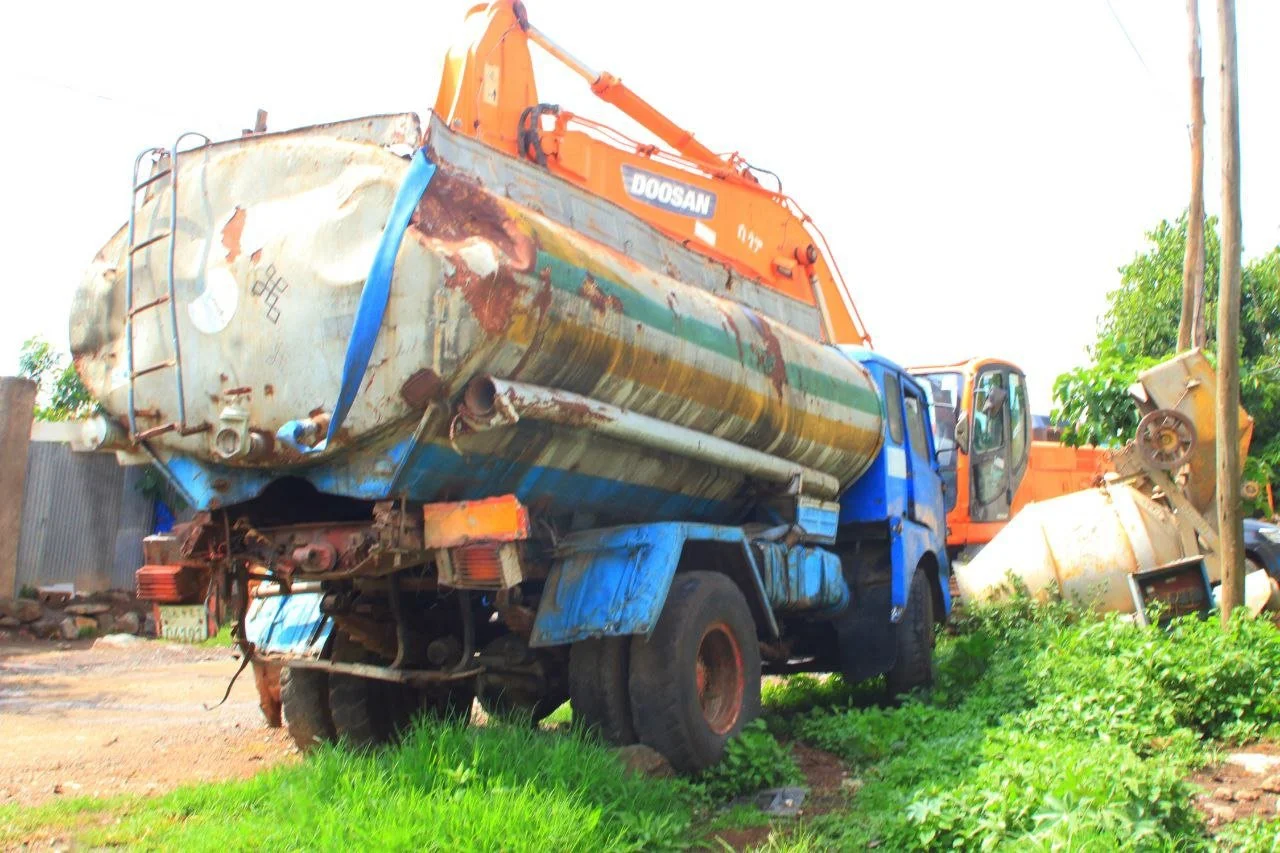 Old, rusty tanker truck with a large orange crane attachment parked on grass and dirt, with green bushes and trees in the background.