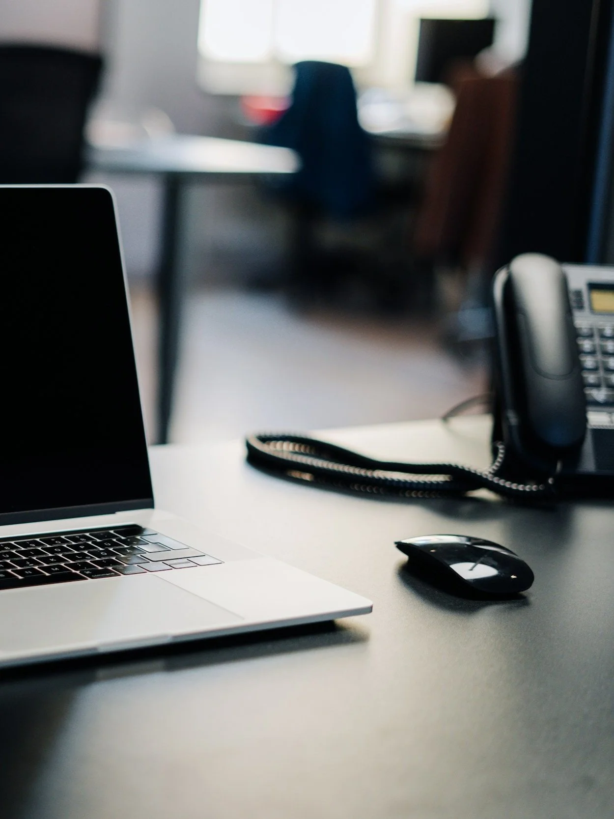 Office desk with a closed laptop, a computer mouse, a telephone, and a coiled cord, with chairs and windows in the background.