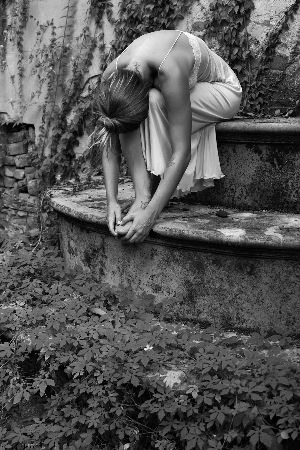 A woman in a flowing dress is sitting on a stone ledge, bending forward. The scene is outdoors, surrounded by overgrown plants and a weathered stone wall.