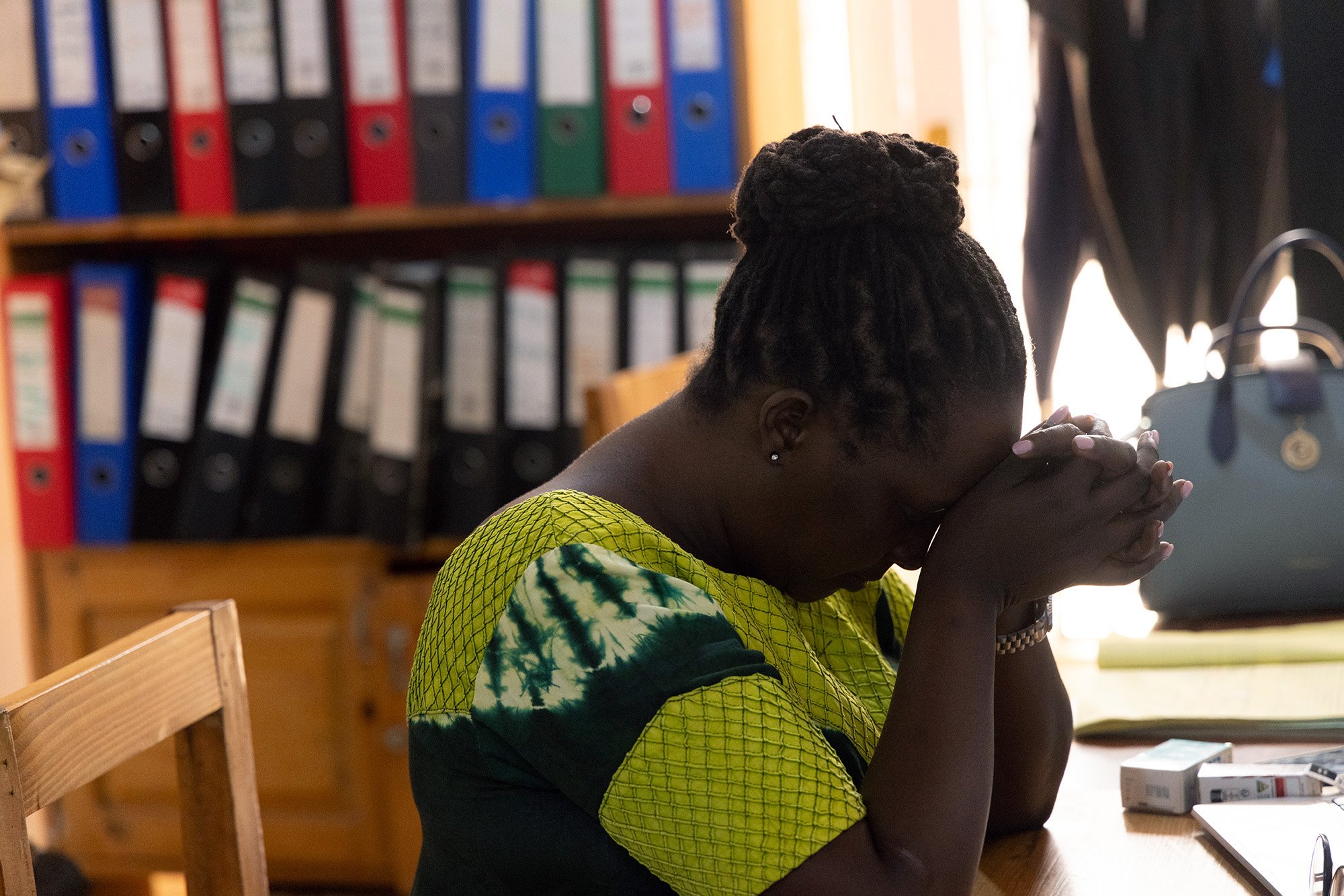 Mary Nyangoma, Project Manager at the Nyamirambo Women’s center, sits at her desk with her head bowed and hands clasped before starting her day on July 5, 2024, in Kigali, Rwanda. 
