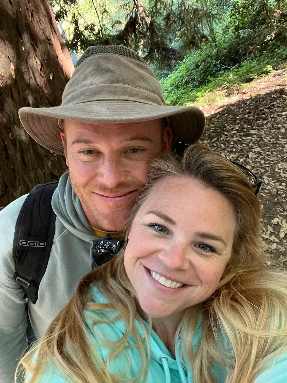 A smiling couple taking a selfie in a wooded outdoor area with trees and fallen leaves in the background.