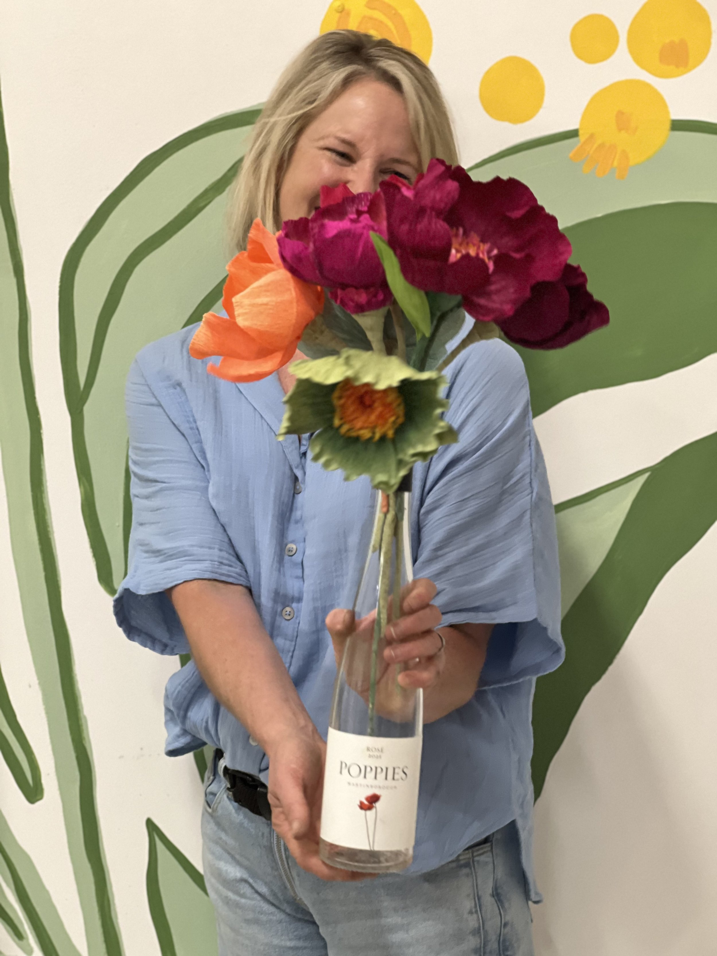 Woman in a light blue shirt holding a glass bottle with colorful artificial poppies in front of a painted floral wall.