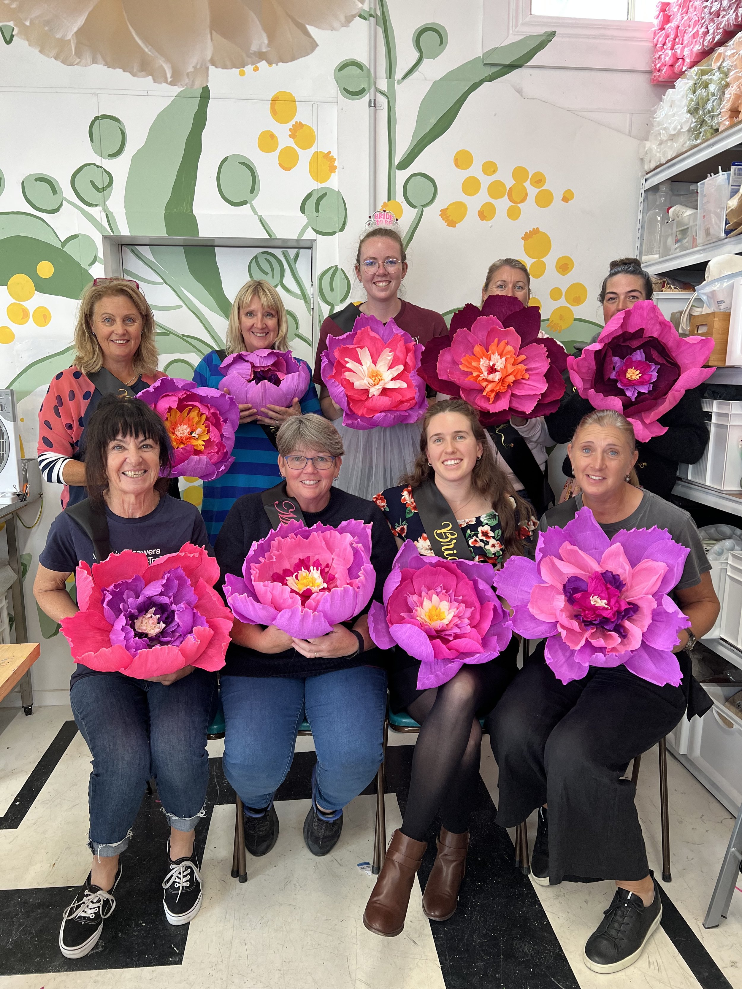A group of ten women at a hen party, holding large artificial pink and purple flowers, celebrating a special occasion with festive sashes. The room has a colorful mural on the wall at Ventana art space in Martinborough..