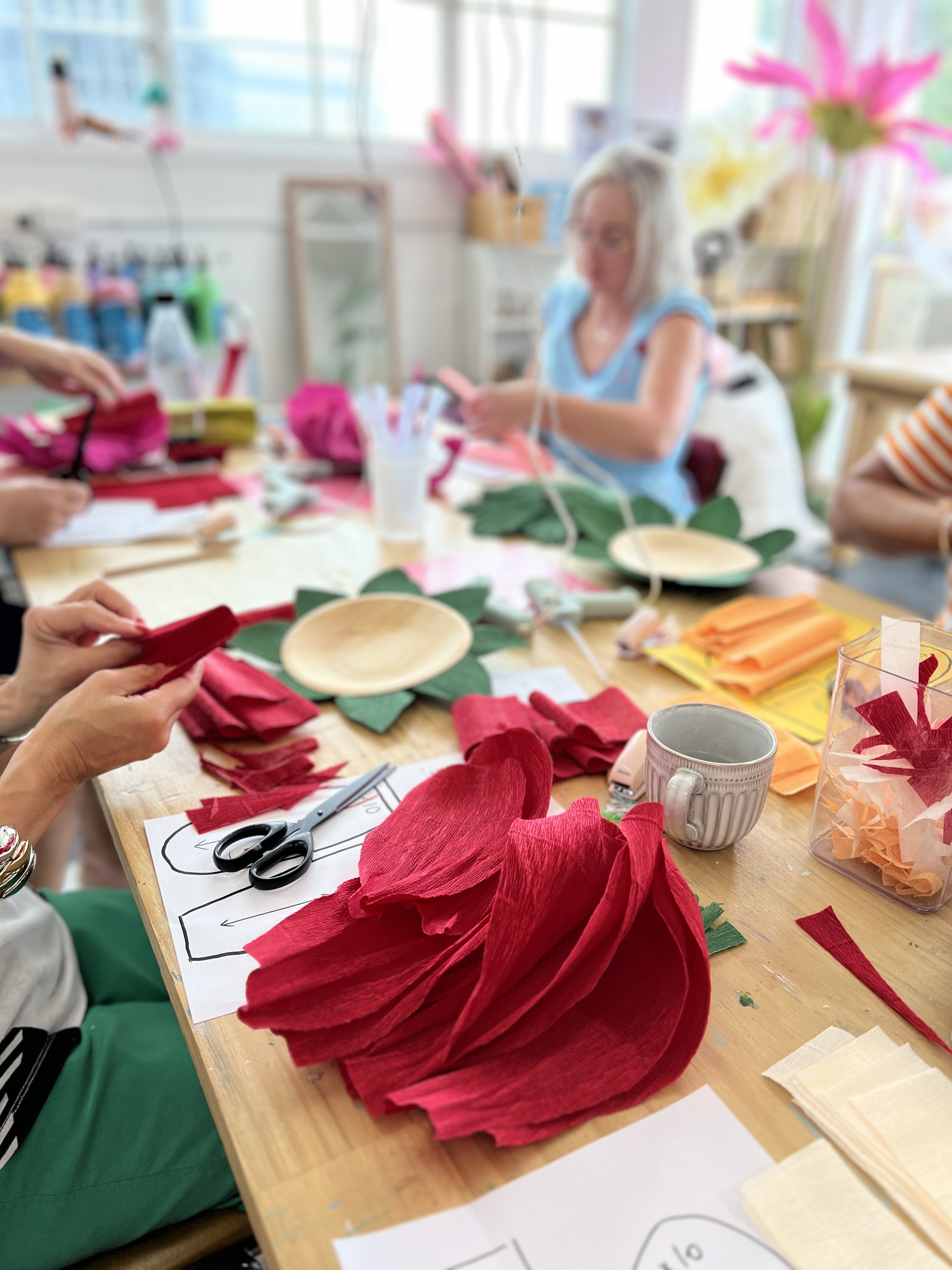 People making paper flowers with red, pink, green, and orange tissue paper and scissors in a craft room.
