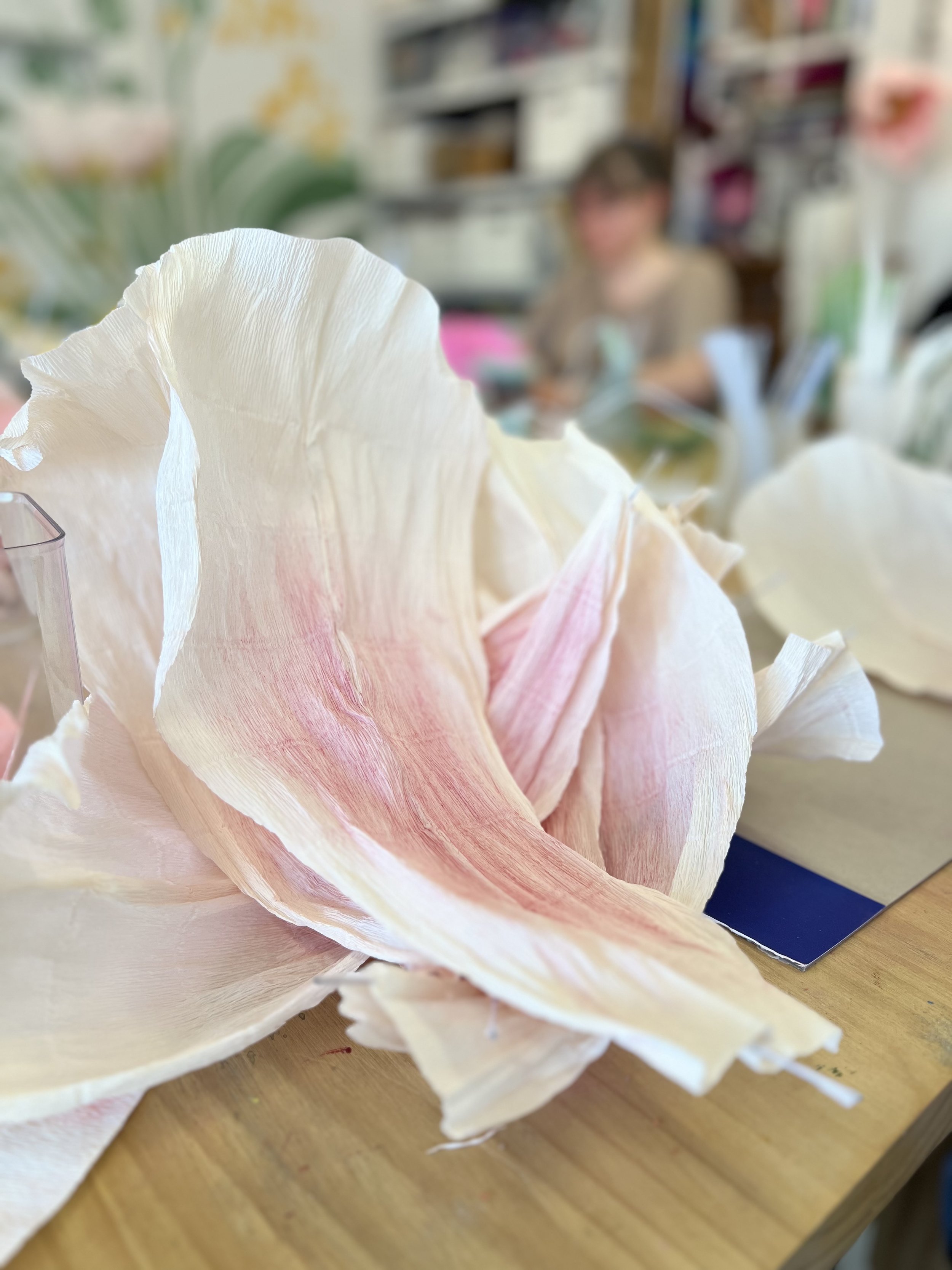 Close-up of a large, pale pink and white artificial flower on a wooden table, with a blurred person and background in a workshop or crafting space.