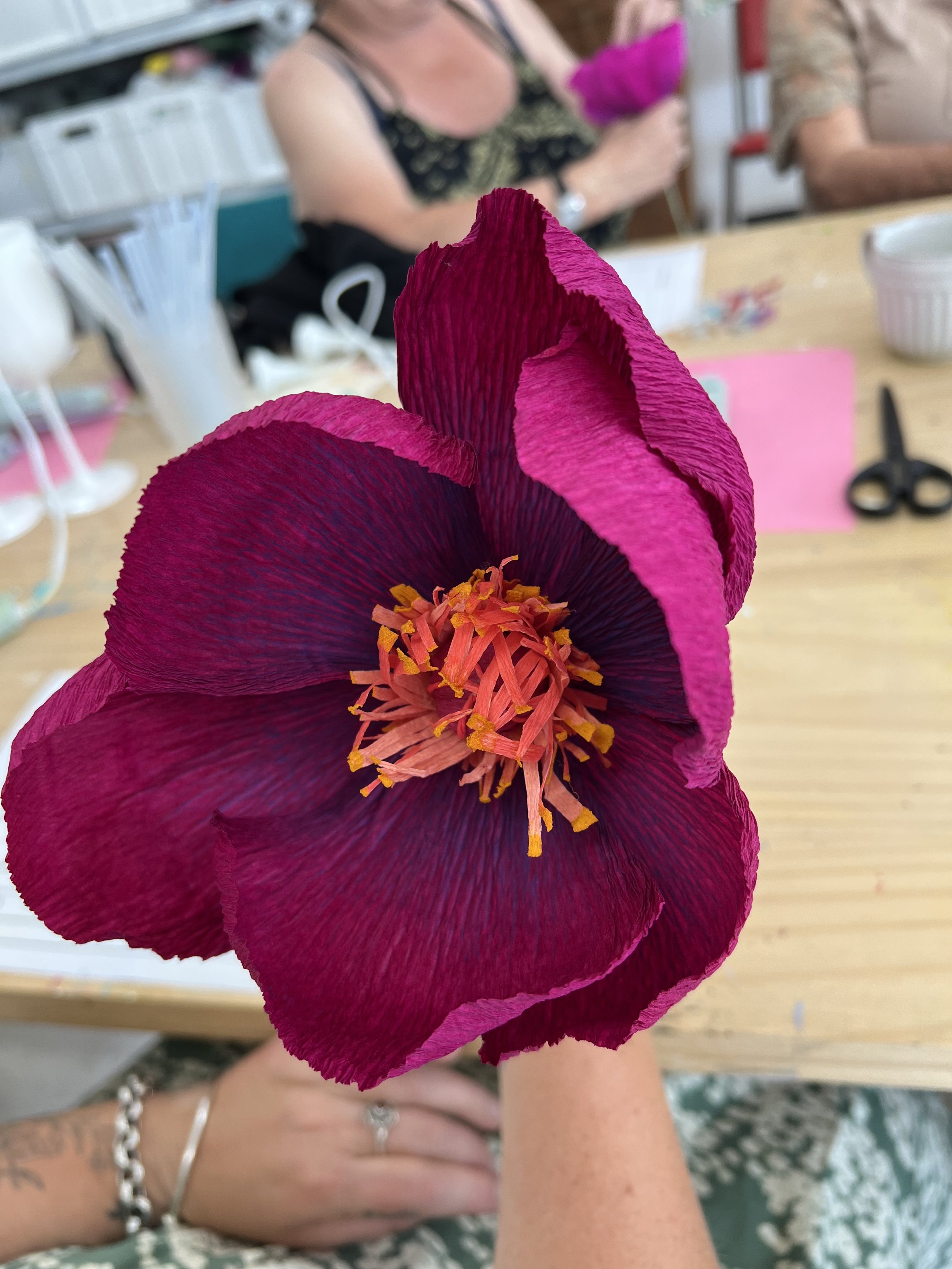 Close-up of a large pink and dark maroon artificial flower with orange and yellow stamens, held by a person with visible tattoos and jewelry, on a table with crafting supplies and people in the background.