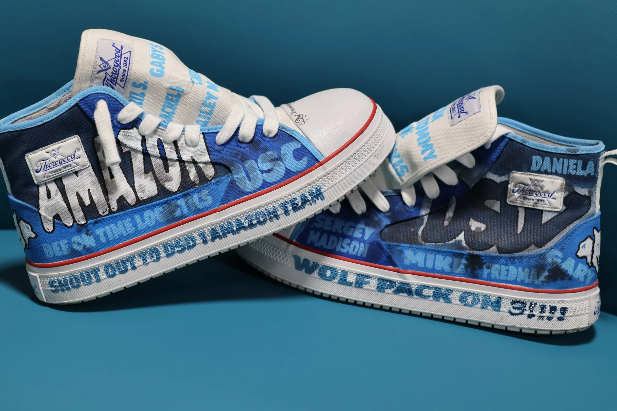 Pair of custom high-top sneakers with blue, white, and black design featuring names and phrases. The left shoe has a white toe cap, white laces, and a white tag with purple text, while the right shoe has a strap with the name Daniel. Both shoes have