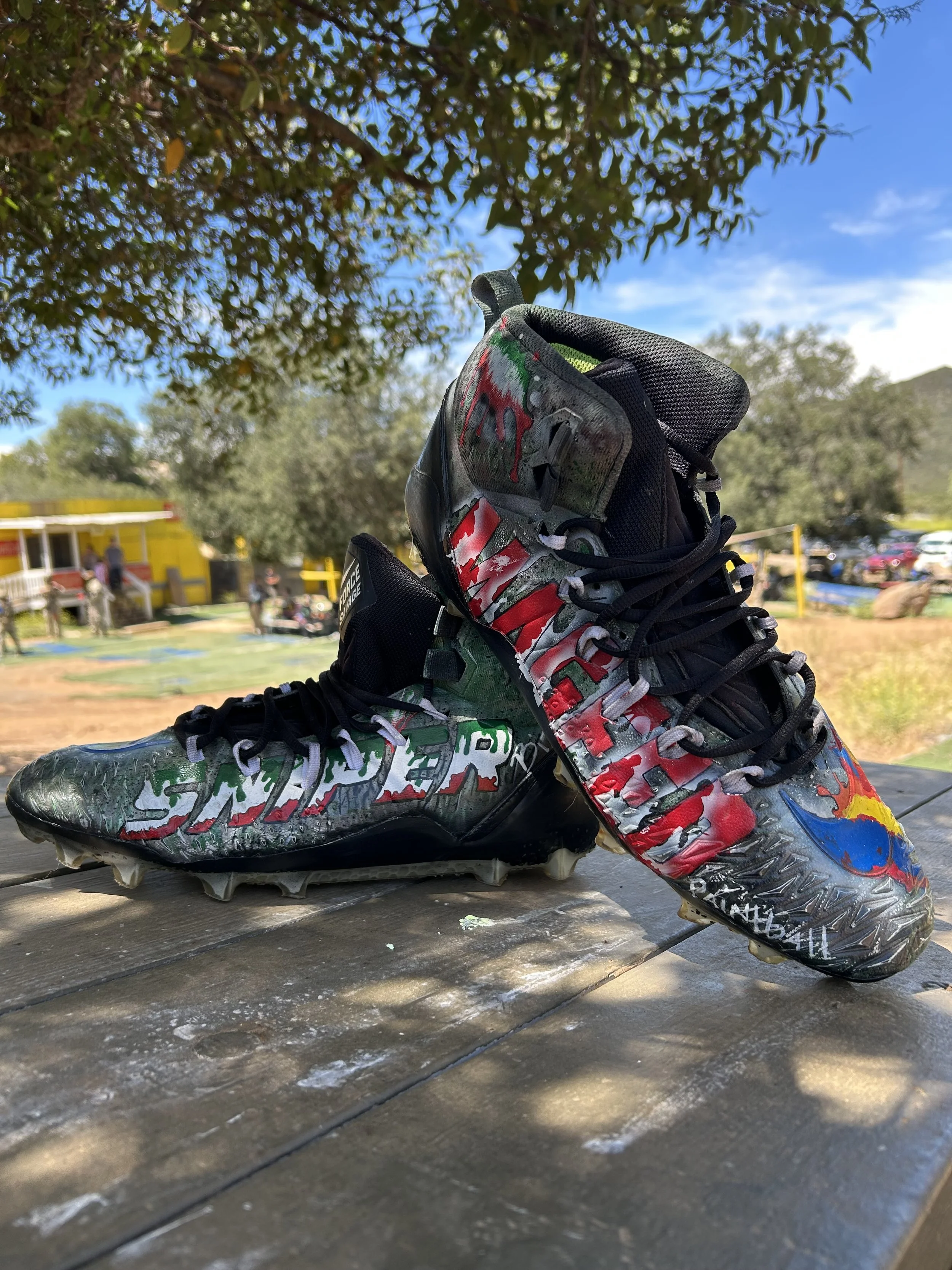 Pair of football cleats with colorful, graffiti-style design sitting on a wooden surface outdoors with trees, grass, and a yellow building in the background.